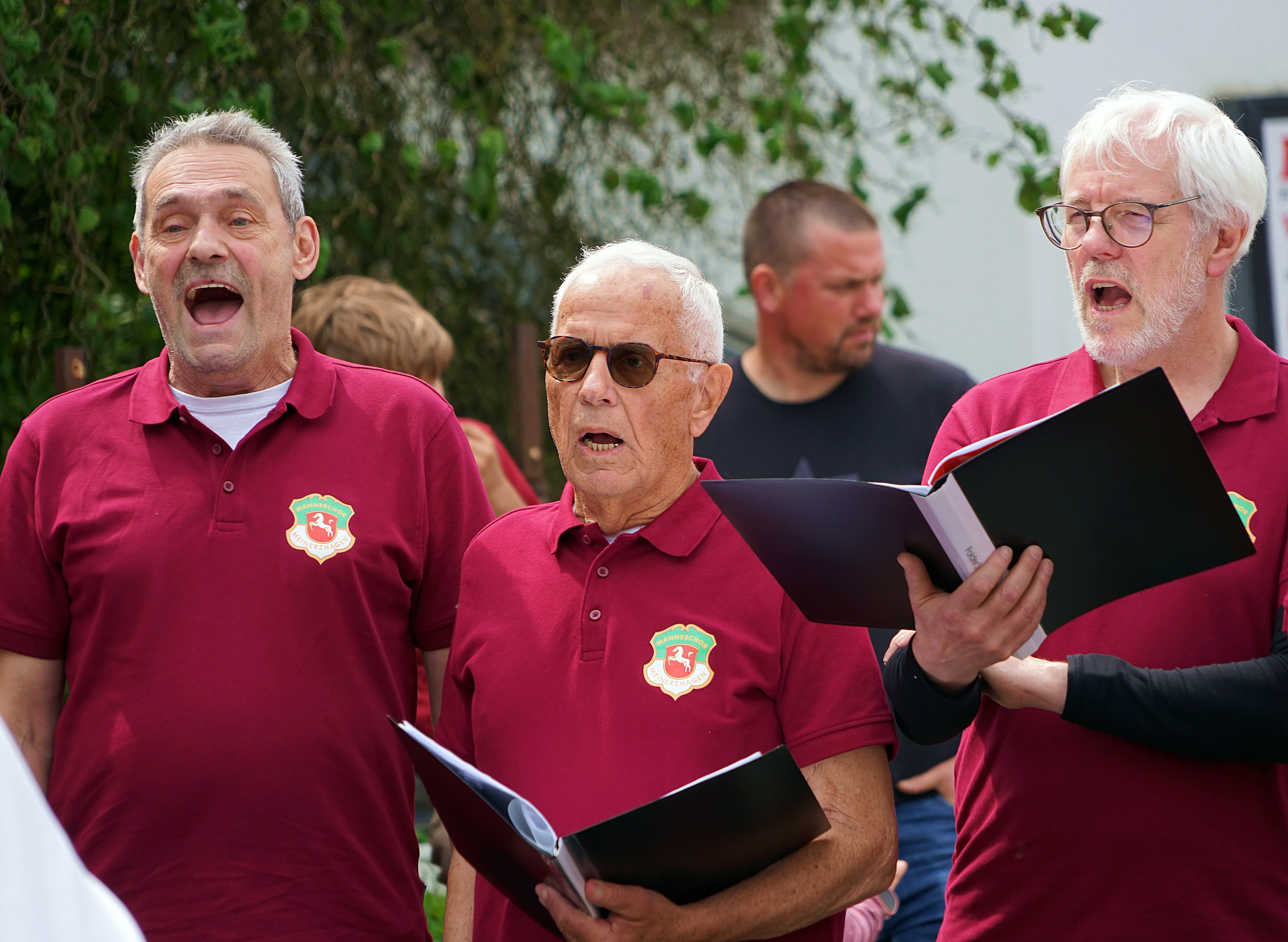 Drei Männer singen im Freien, jeder hält Notenblätter in der Hand. Sie tragen rote Poloshirts mit einem Wappen darauf.