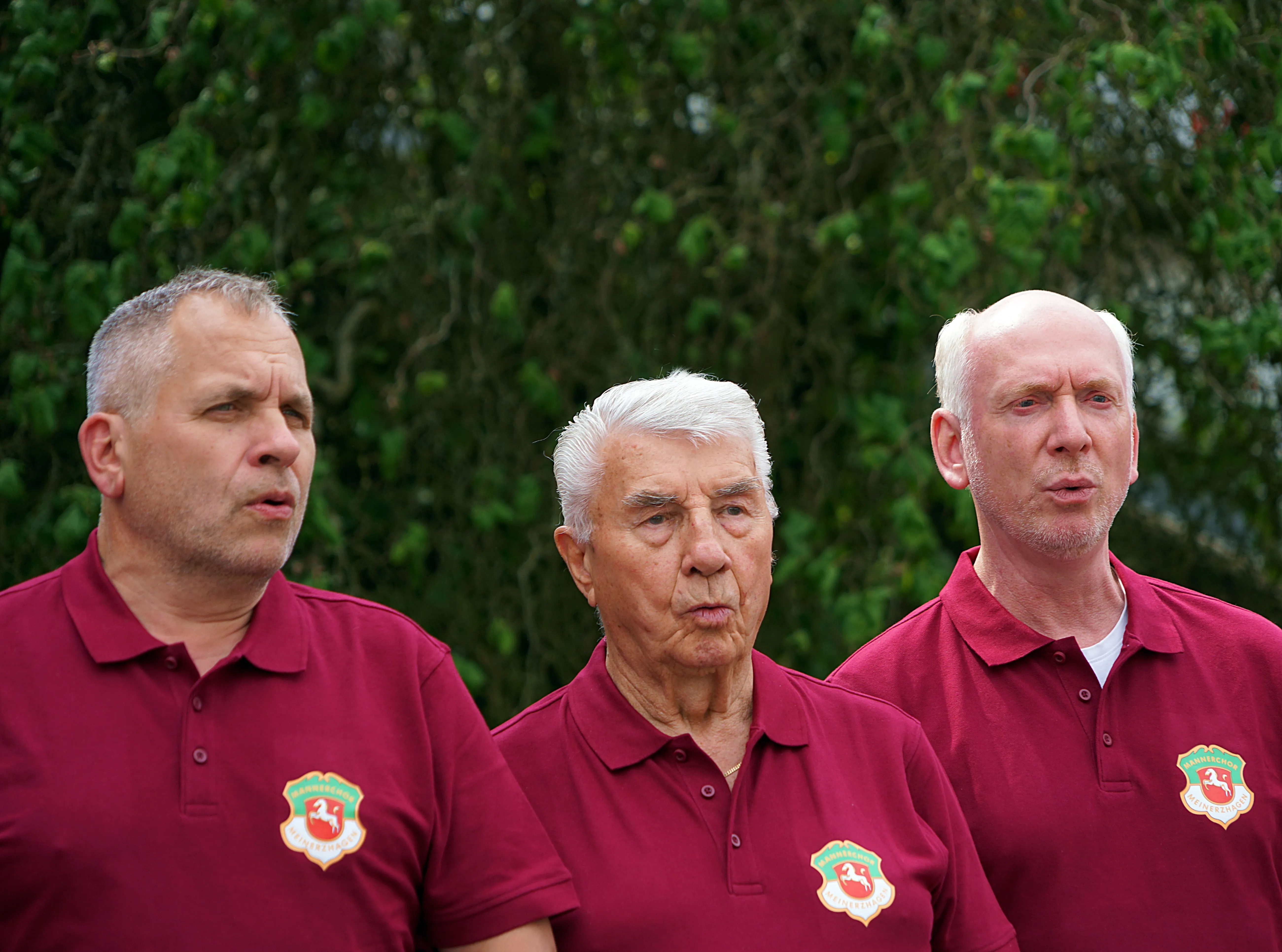 Drei Männer mittleren und höheren Alters mit roten Poloshirts und einem Wappen auf der Brust schauen ernst in die Kamera.