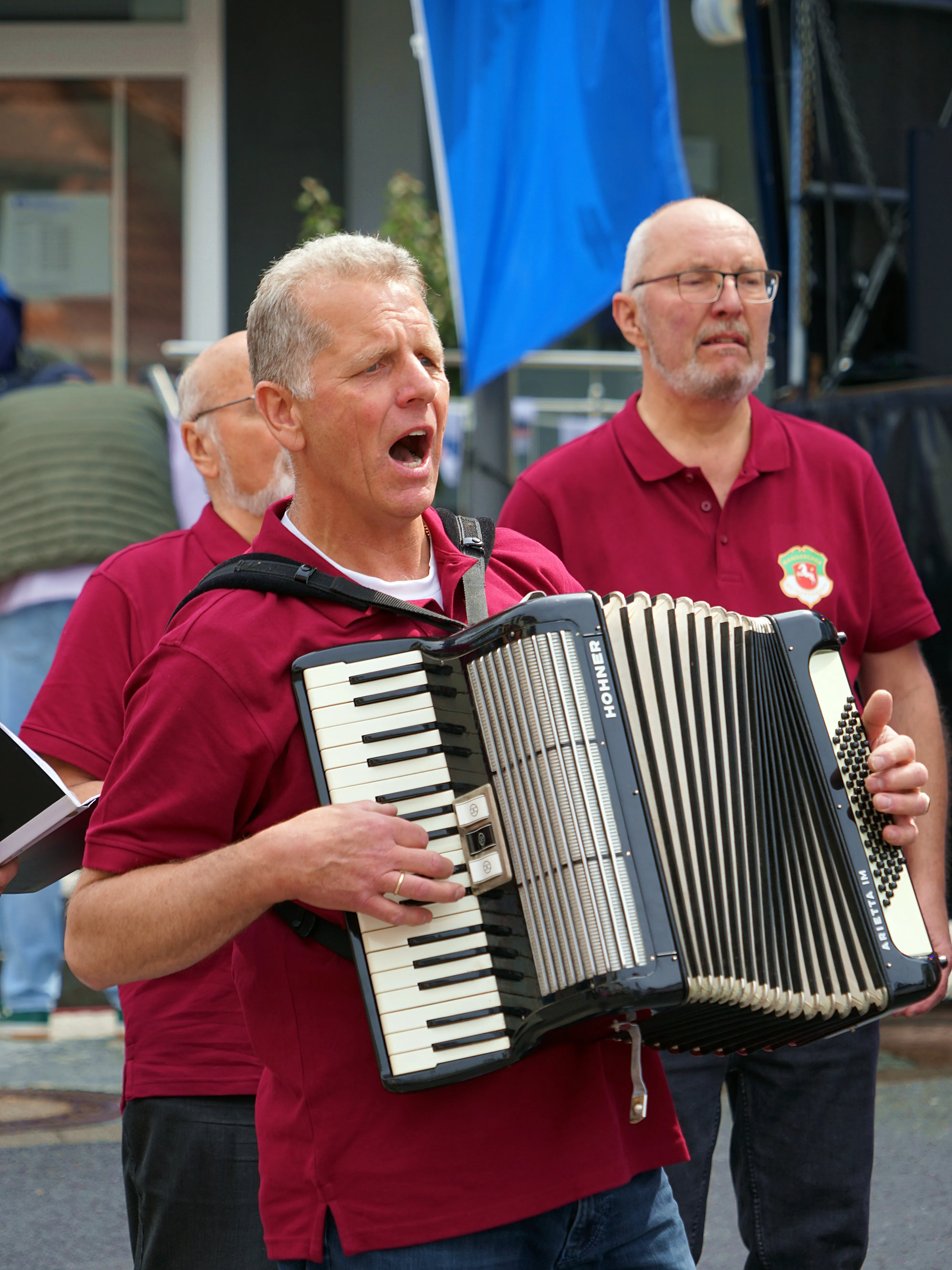 Ein Mann spielt Akkordeon, während er singt, umgeben von zwei weiteren Männern im Hintergrund. Alle tragen rote Poloshirts.