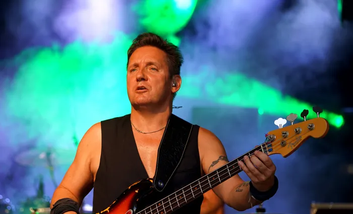 Ein Mann spielt auf einem Bass vor einer Bühne mit grün-blauem Bühnenlicht. Er trägt ein schwarzes Tanktop und hat einen Armverband.
