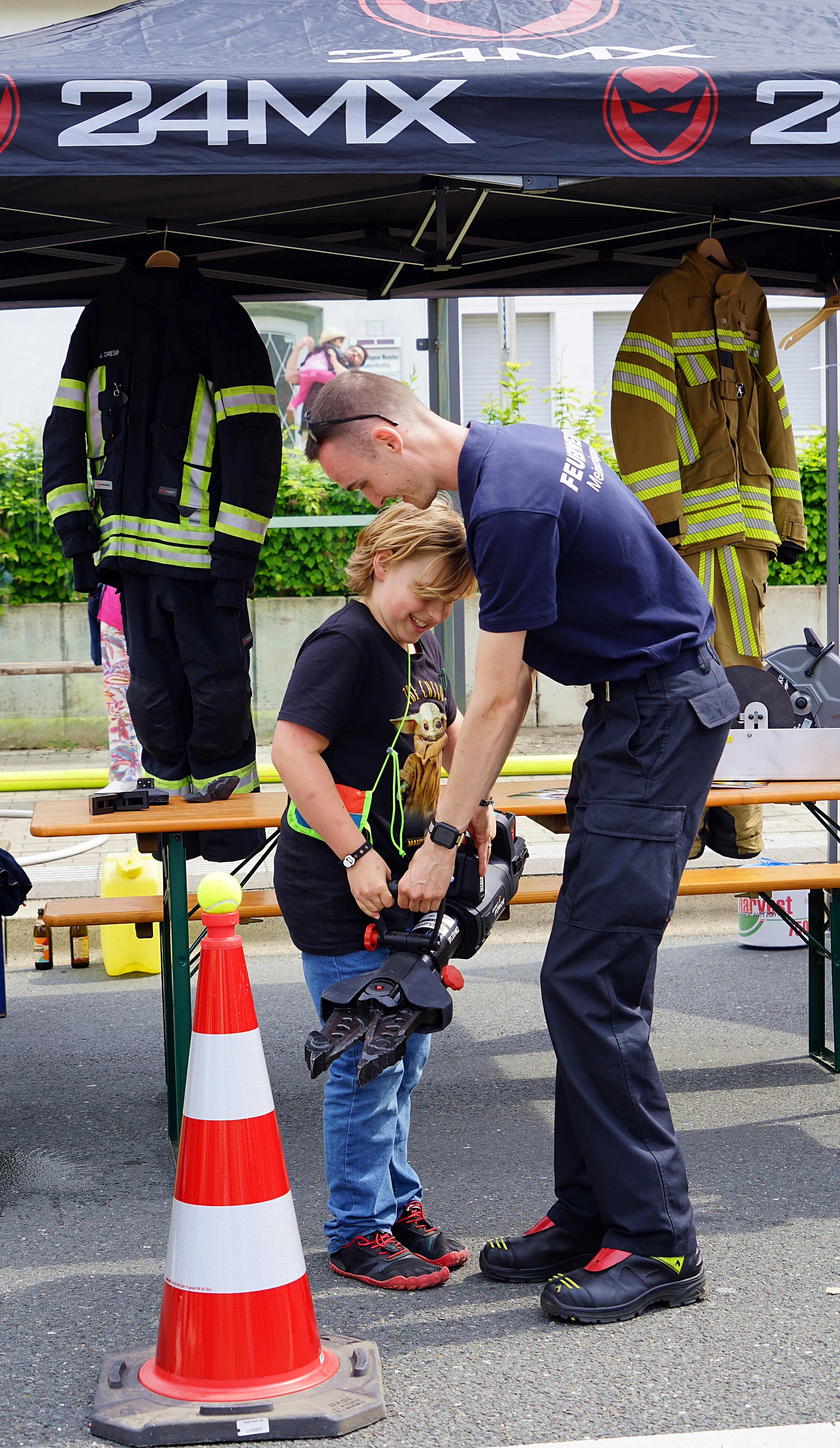 Ein Feuerwehrmann hilft einem Kind, einen Helm aufzusetzen. Im Hintergrund hängen weitere Feuerwehruniformen unter einem Pavillon.