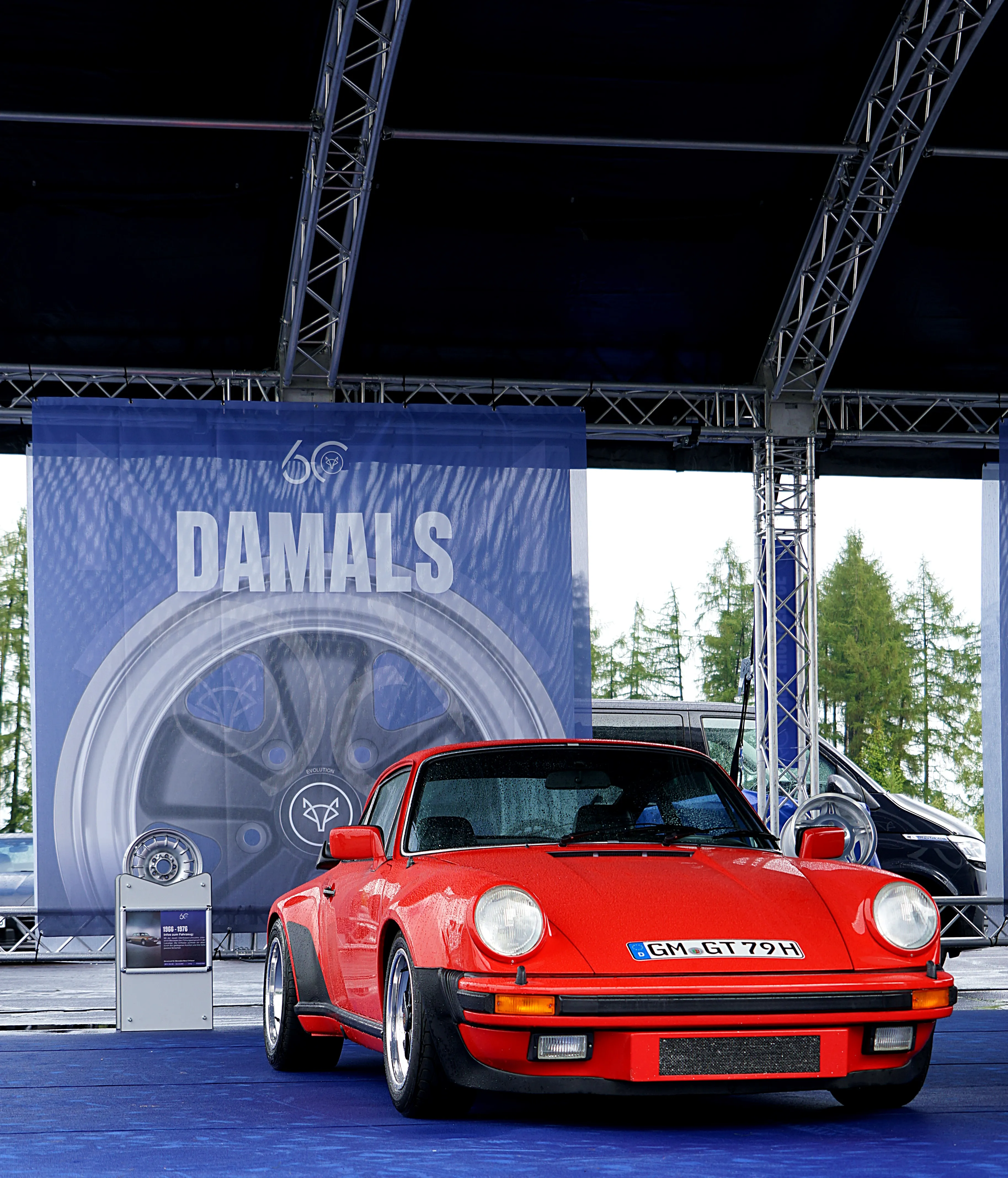 Roter Porsche 911 steht vor einer blauen Wand mit dem Schriftzug „DAMALS“ und dem Logo einer Fuchsfelge. Im Hintergrund sind weitere Fahrzeuge und Bäume zu sehen.
