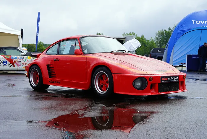 Roter Porsche 935 mit Fuchsfelgen steht auf nasser Fahrbahn, Spiegelung im Asphalt. Im Hintergrund ein Zelt und weitere Fahrzeuge.