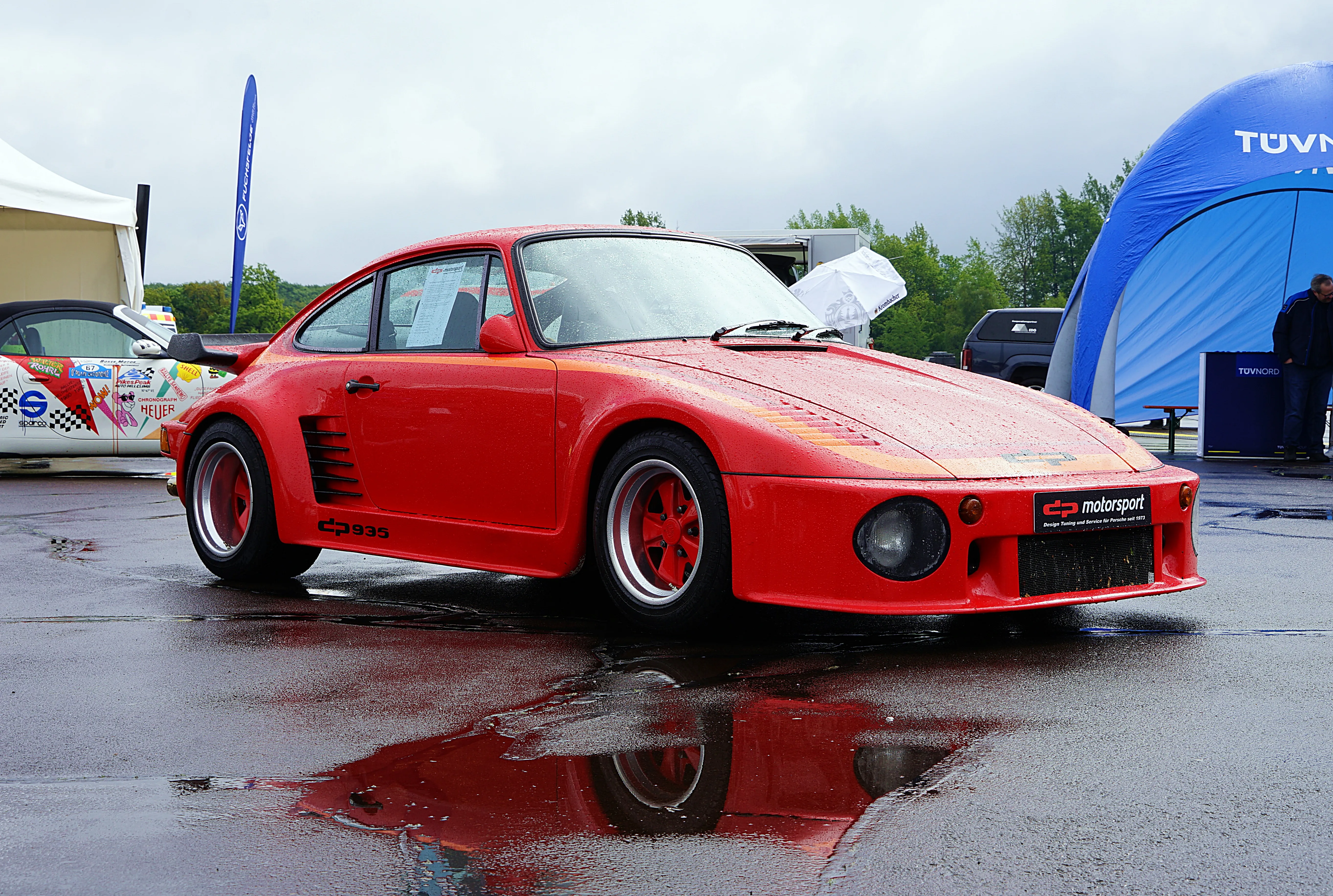 Roter Porsche 935 mit Fuchsfelgen steht auf nasser Fahrbahn, Spiegelung im Asphalt. Im Hintergrund ein Zelt und weitere Fahrzeuge.