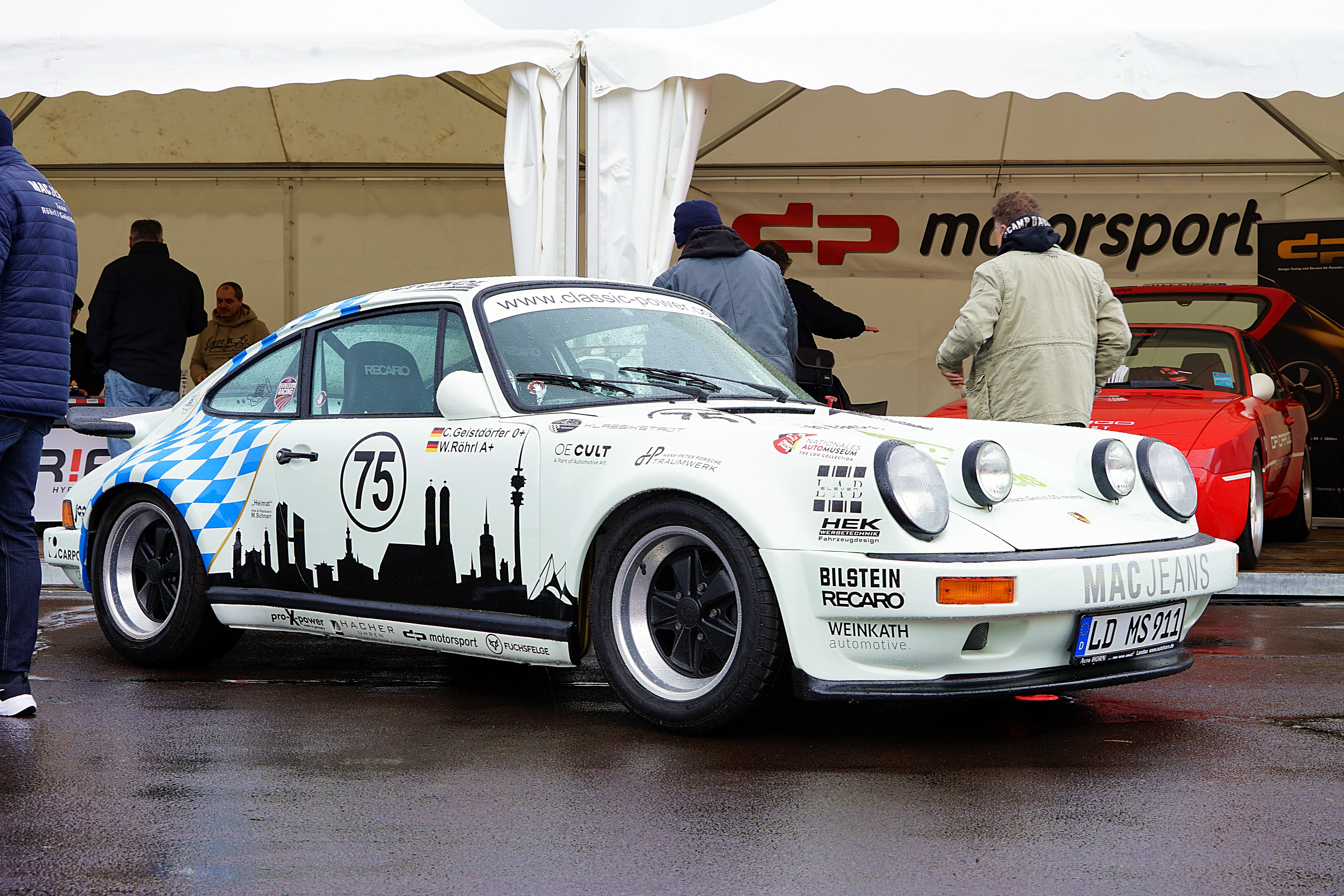 Ein dunkelblauer Porsche 911 mit Startnummer 75 und Skyline-Motiv steht in einer Garage, umgeben von Zuschauern und einem roten Sportwagen.