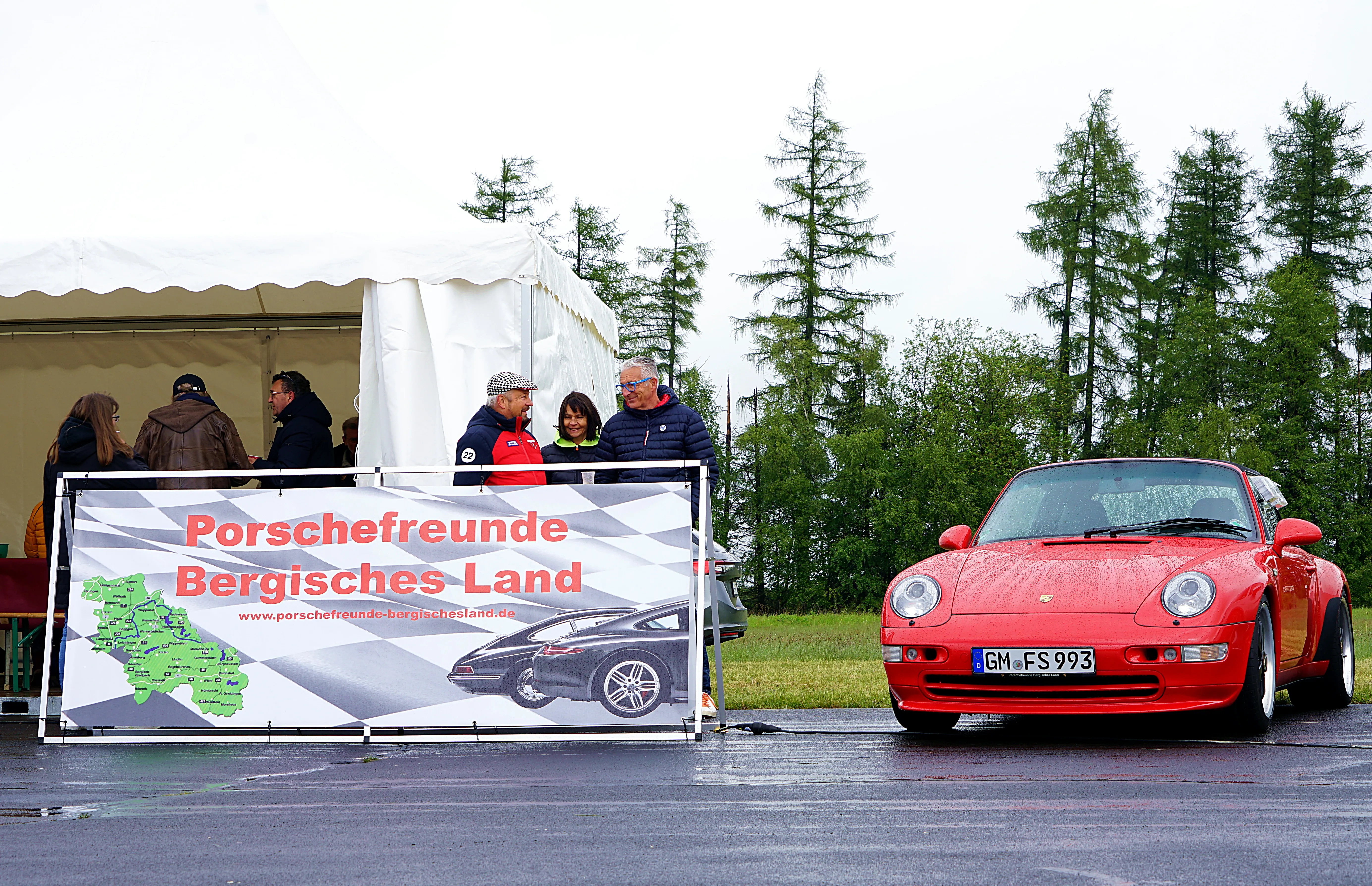 Roter Porsche 911 steht vor einem Zelt mit dem Banner „Porschefreunde Bergisches Land“. Im Hintergrund stehen weitere Personen und Bäume.