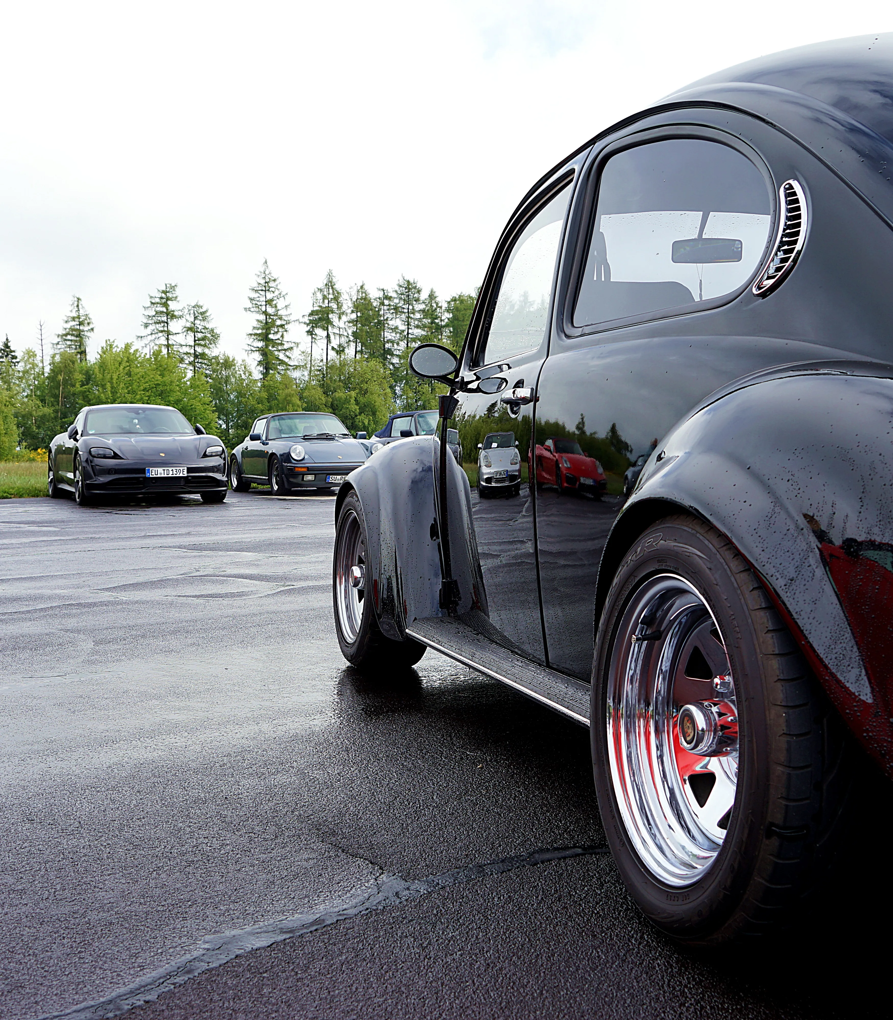 Ein schwarzer VW Käfer mit glänzenden Fuchsfelgen steht im Vordergrund, während im Hintergrund verschiedene Porsche-Modelle auf einem Parkplatz stehen.