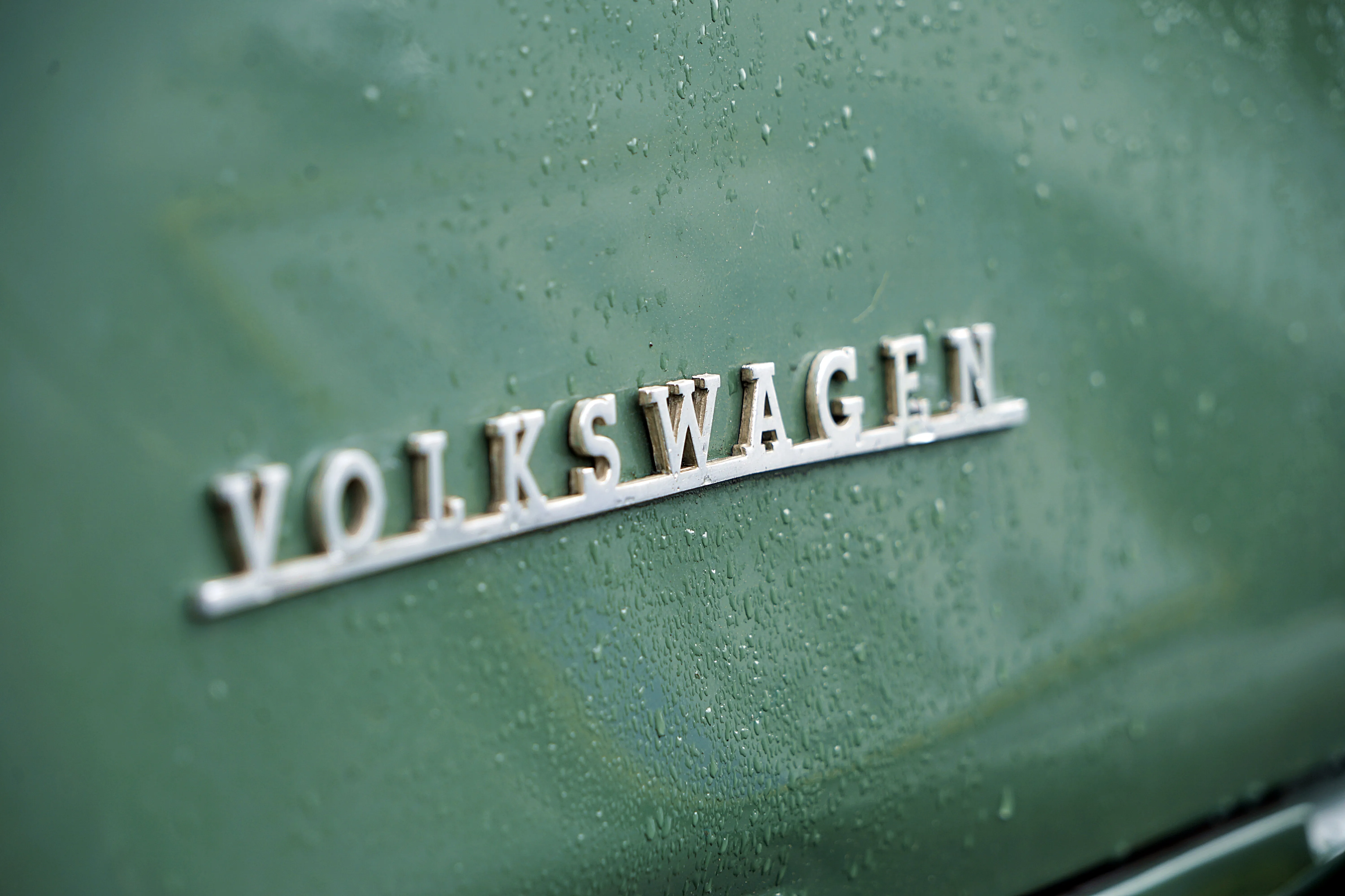 Chromglänzendes Volkswagen-Emblem auf einem hellgrünen Fahrzeug, bedeckt mit Wassertropfen.