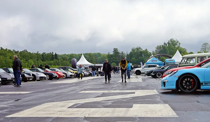 Eine Reihe von Sportwagen steht auf einem abgesperrten Asphaltplatz, während Besucher zwischen den Fahrzeugen herumlaufen. Im Hintergrund sind weiße Zelte und Bäume zu sehen.