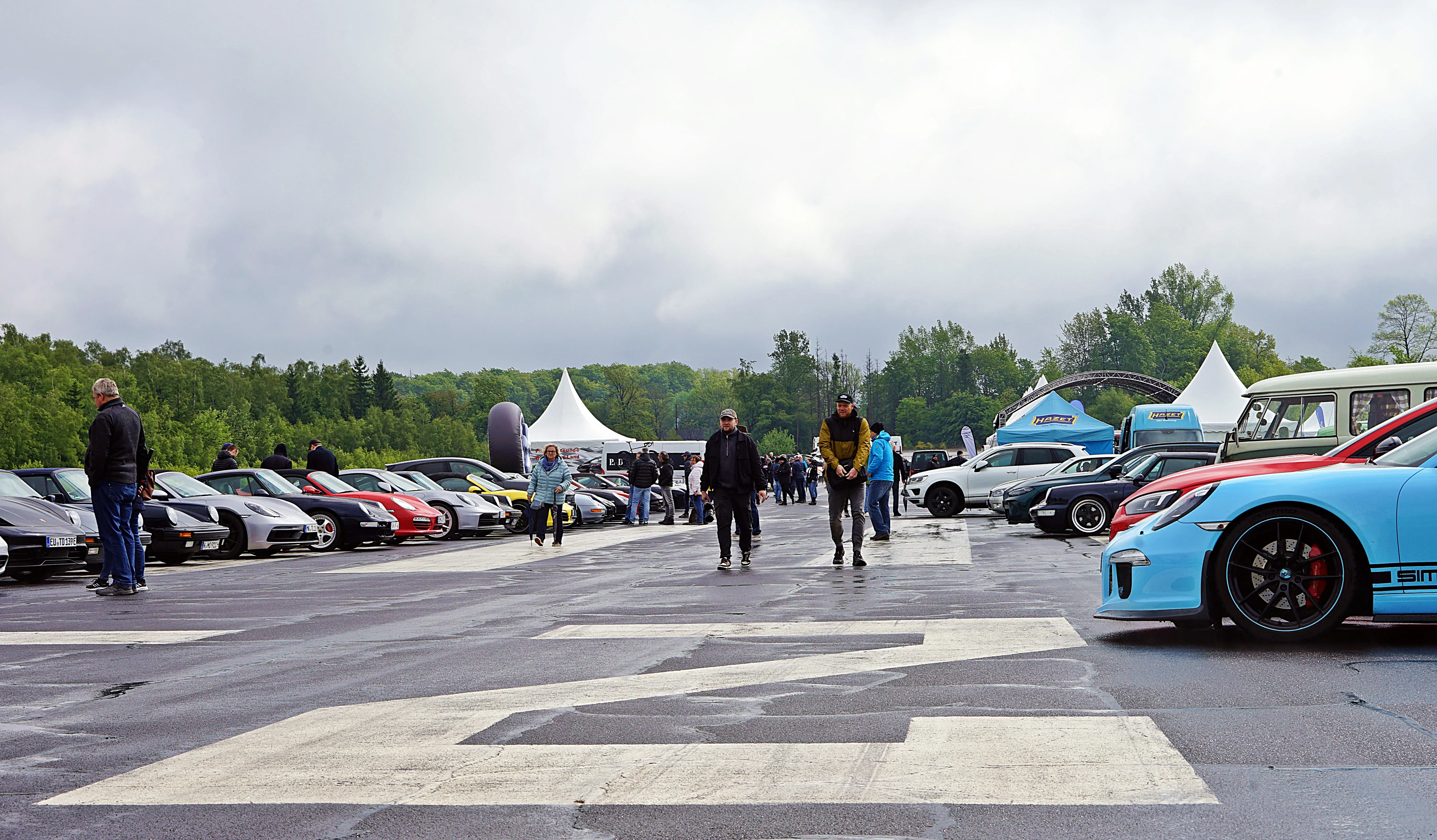 Eine Reihe von Sportwagen steht auf einem abgesperrten Asphaltplatz, während Besucher zwischen den Fahrzeugen herumlaufen. Im Hintergrund sind weiße Zelte und Bäume zu sehen.