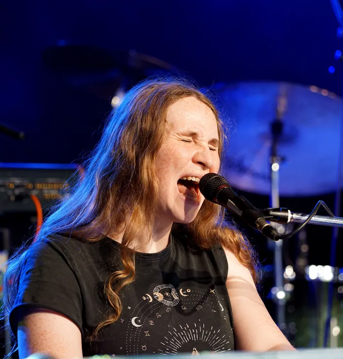 Ein rothaariger Mann mit langen Haaren singt in ein Mikrofon. Er trägt ein schwarzes T-Shirt mit einem grafischen Muster aus Sternen und Monden und spielt auf einer elektronischen Orgel.