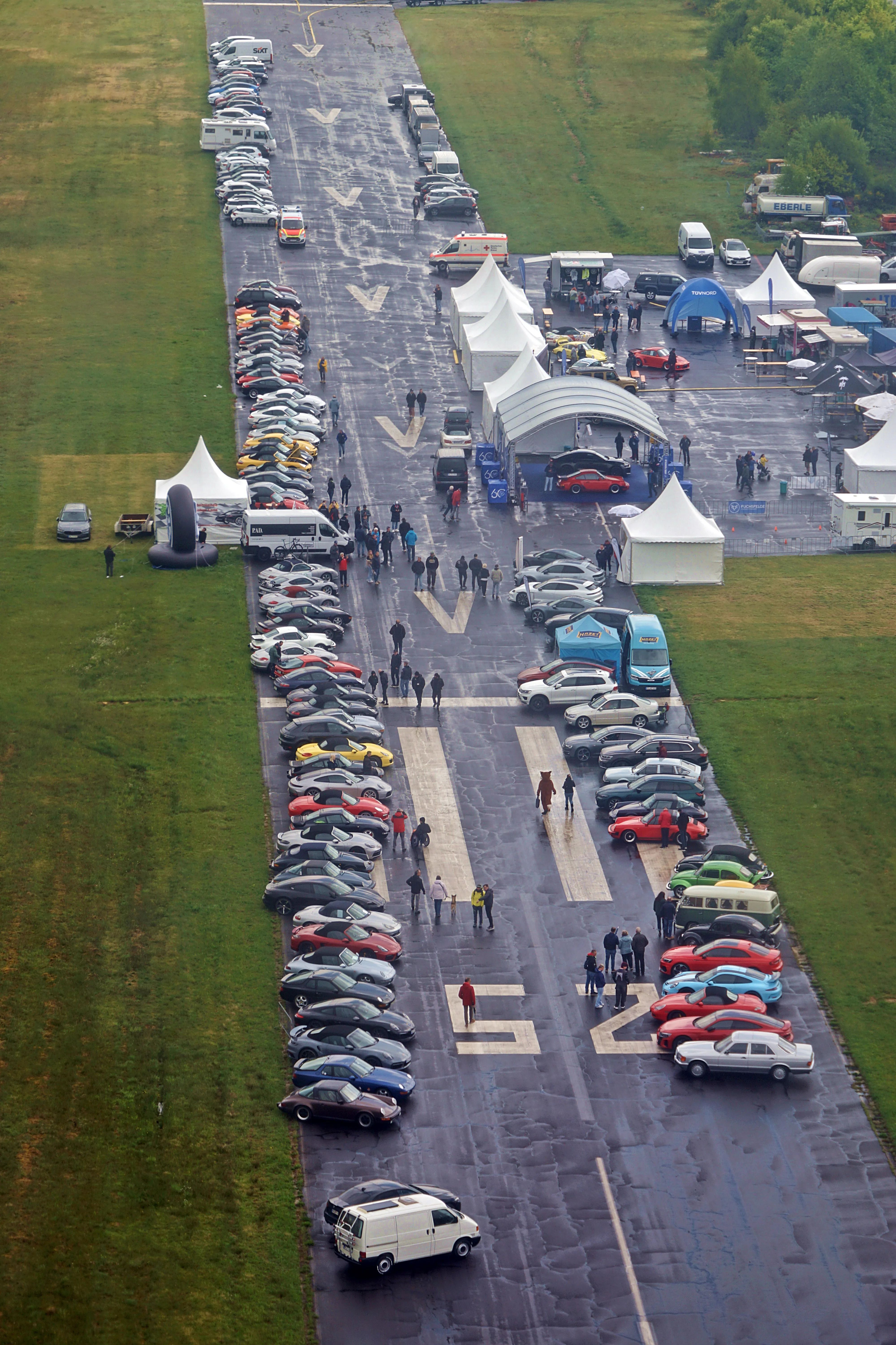 Eine lange Reihe von Autos steht auf einem nassen Asphaltstreifen, umgeben von grünen Wiesen und mehreren weißen Zelten. Im Hintergrund sind weitere Fahrzeuge und Menschen zu sehen.