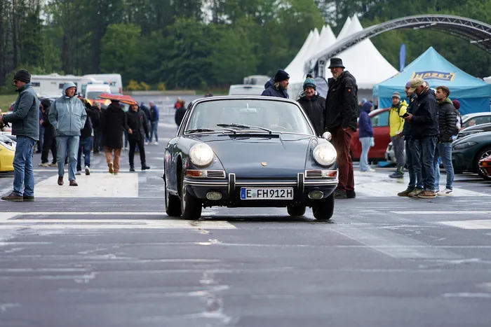 Grauer Porsche 911 fährt auf einem Parkplatz vor einer Menschenmenge und mehreren Zelten. Im Hintergrund ist ein Werbebanner für Hazet zu sehen.