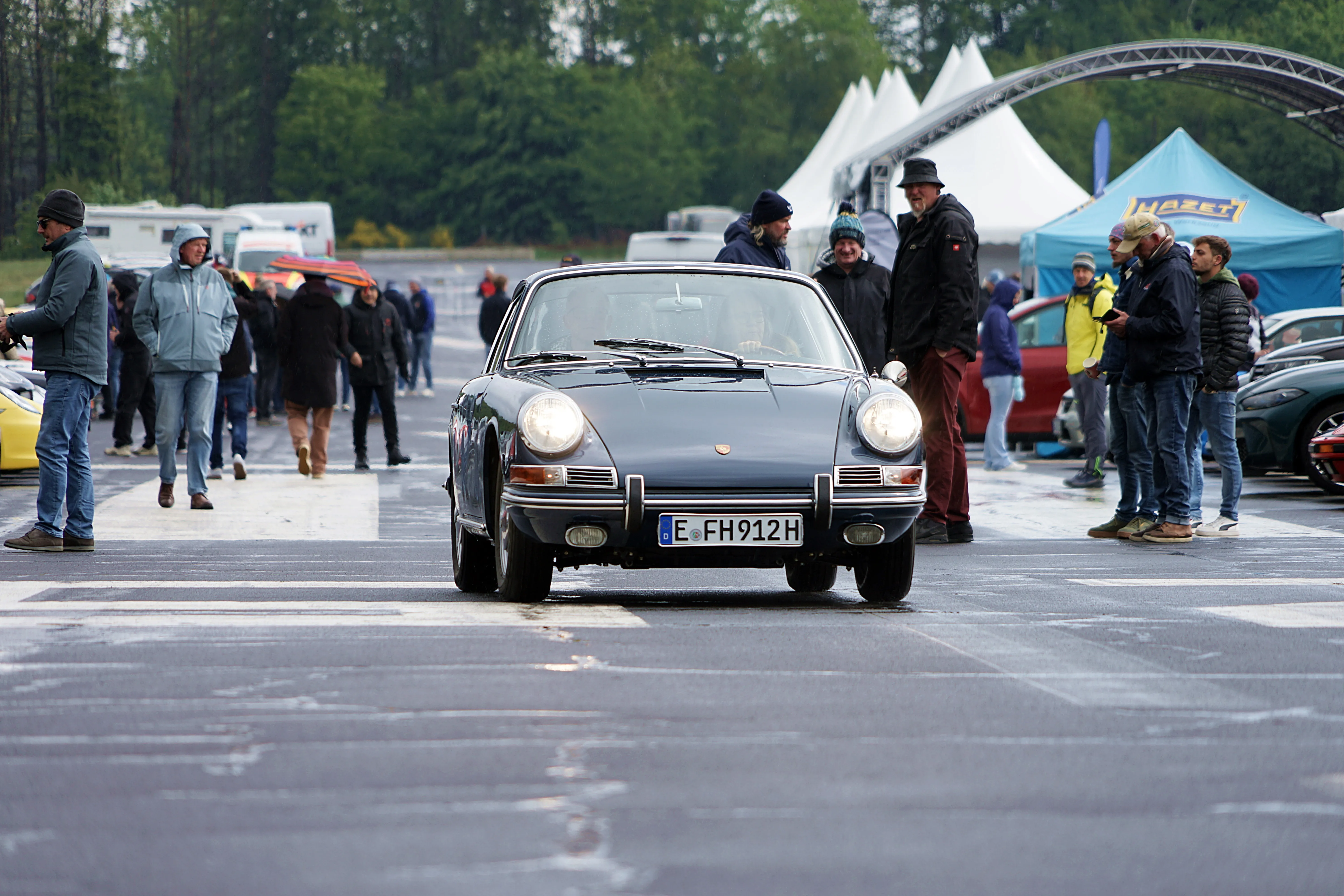 Grauer Porsche 911 fährt auf einem Parkplatz vor einer Menschenmenge und mehreren Zelten. Im Hintergrund ist ein Werbebanner für Hazet zu sehen.