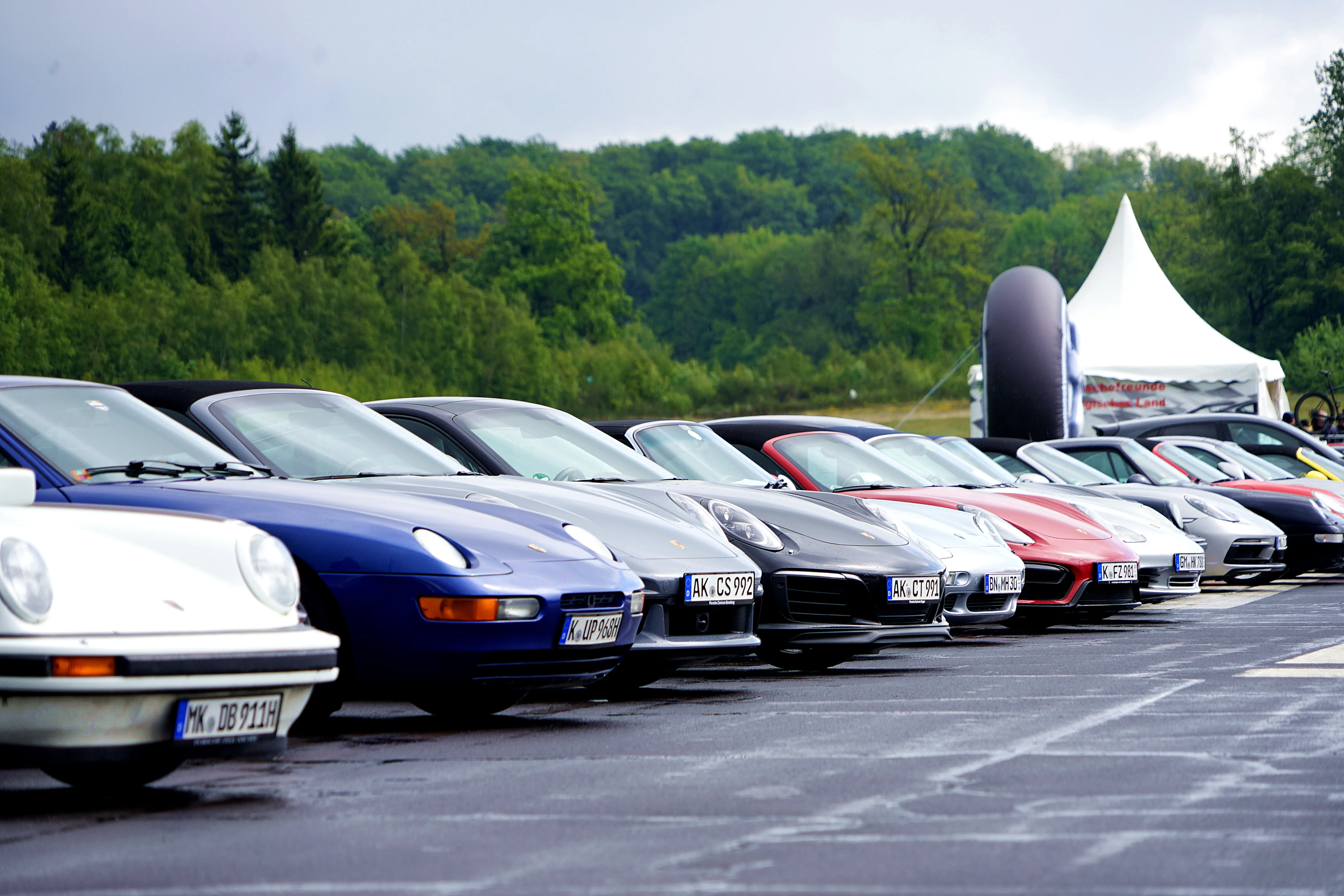 Reihe verschiedener Porsche-Modelle auf einem Parkplatz, darunter ein klassischer Elfer in Hellgelb und ein moderner 911 in Rot. Im Hintergrund Bäume und ein Zelt.