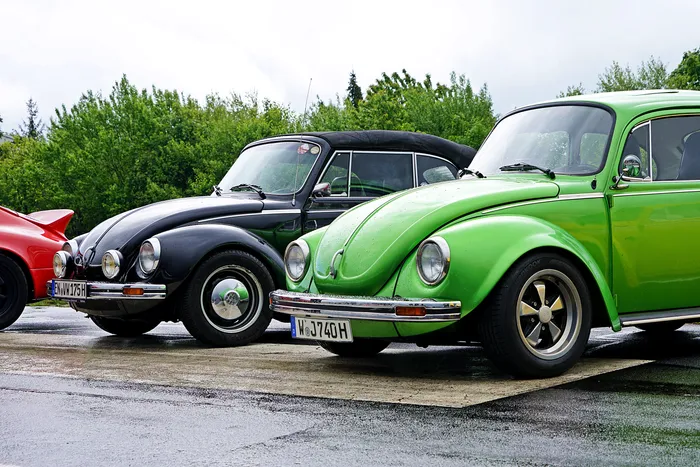 Drei Oldtimer-VW-Käfer in Schwarz, Grün und Rot stehen auf einem Parkplatz vor grüner Vegetation. Die grünen und schwarzen Fahrzeuge haben Fuchsfelgen.