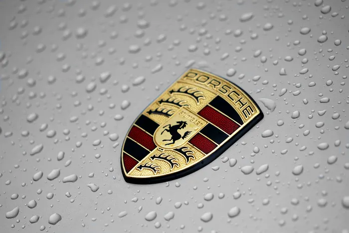 Porsche-Emblem auf einem Fahrzeug, umgeben von Wassertropfen. Das Logo zeigt ein Wappen mit einem Pferd und der Schrift 'Porsche' sowie 'Stuttgart'.