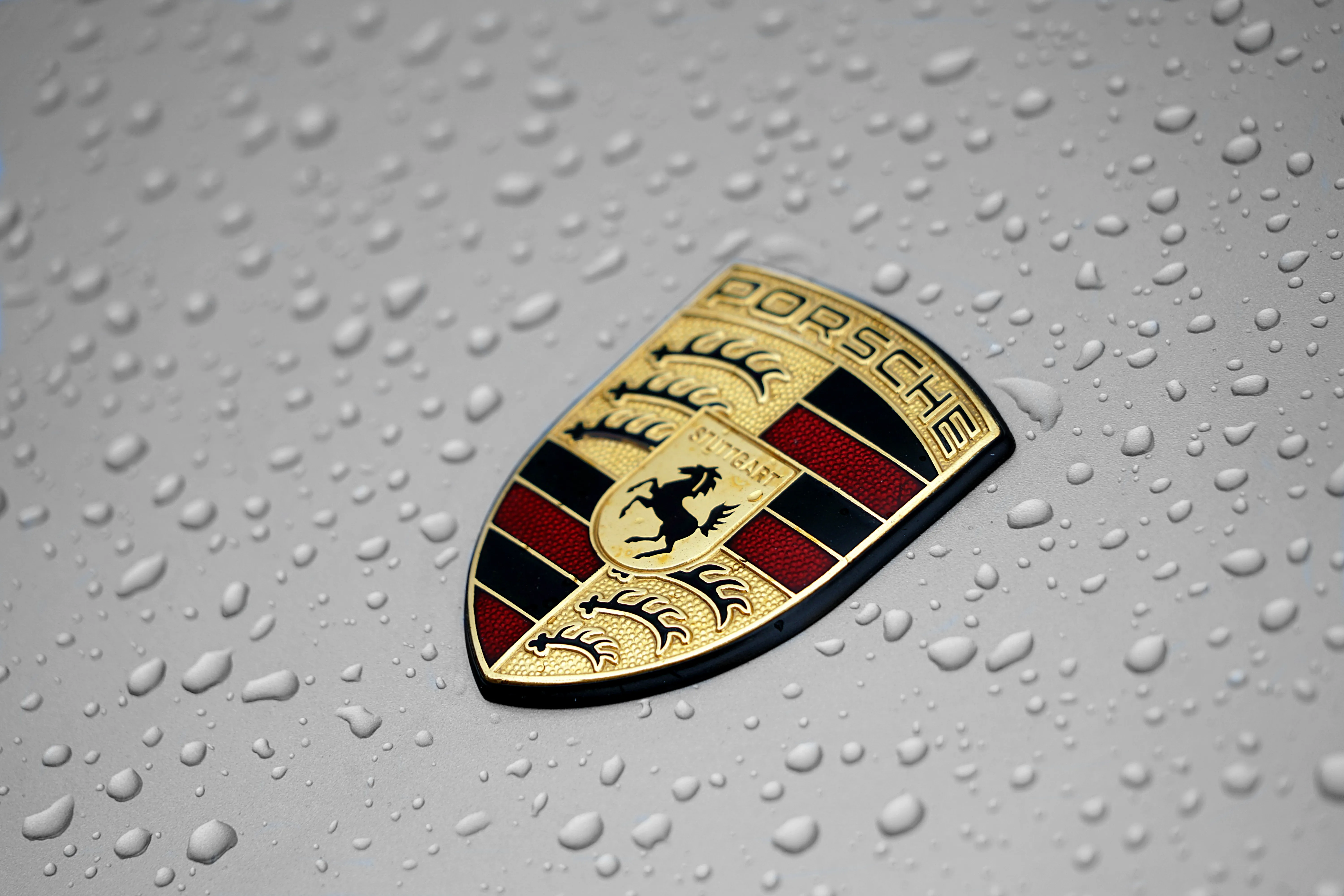 Porsche-Emblem auf einem Fahrzeug, umgeben von Wassertropfen. Das Logo zeigt ein Wappen mit einem Pferd und der Schrift 'Porsche' sowie 'Stuttgart'.