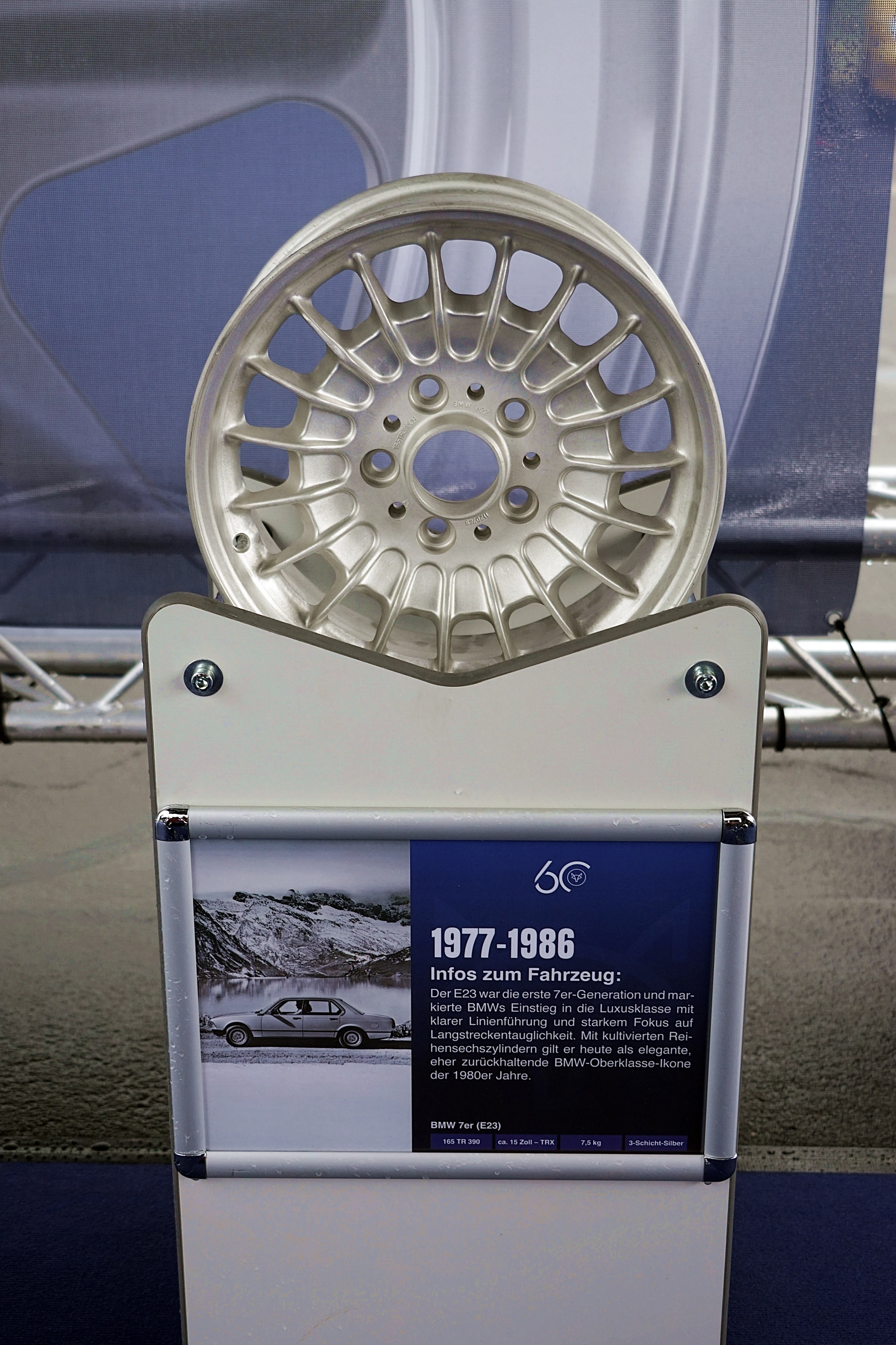 Nahaufnahme einer klassischen Fuchsfelge mit fünf Speichen, präsentiert neben einem Informationsstand mit einem historischen BMW 7er (E23) und technischen Daten.