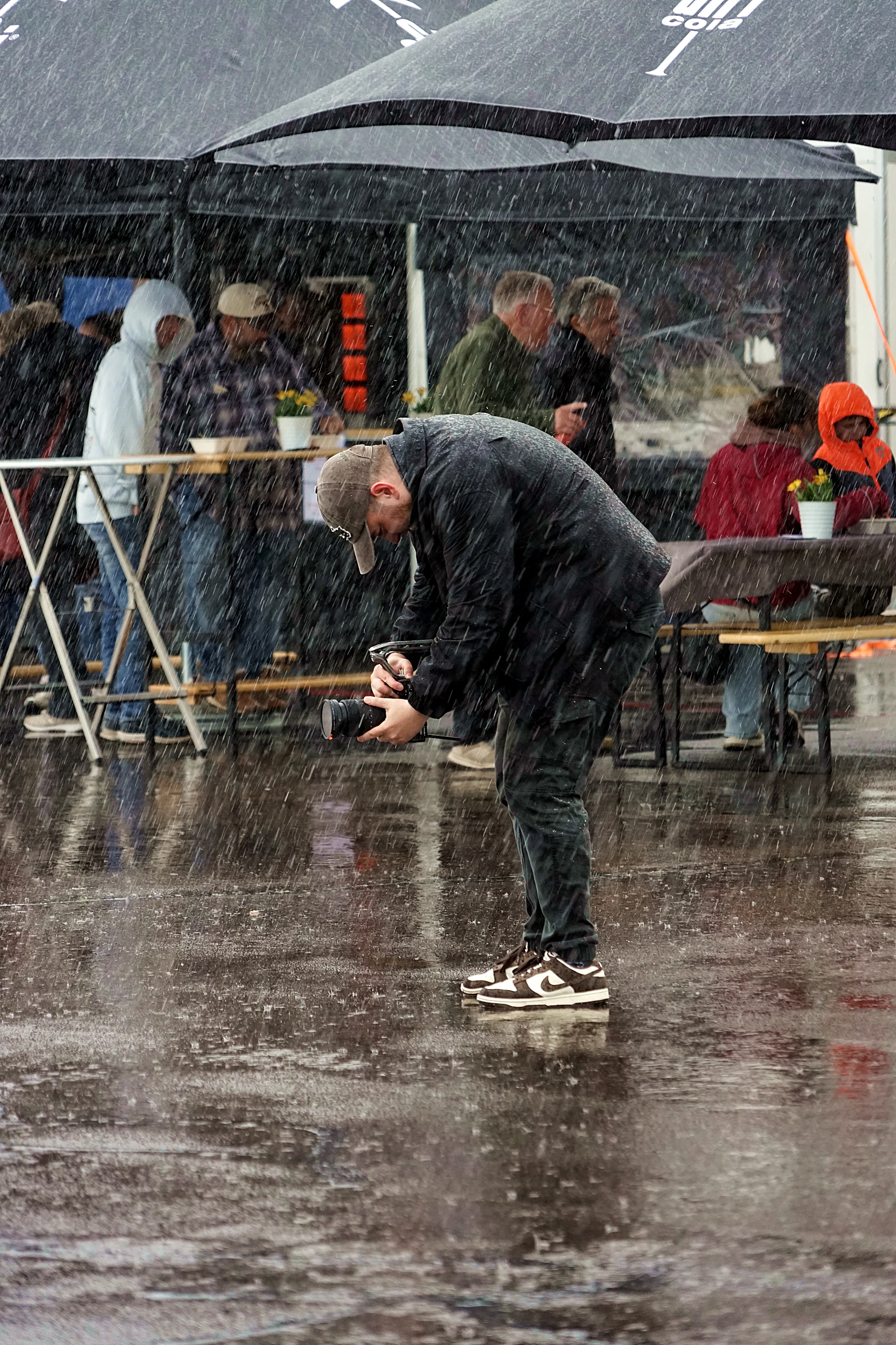 Ein Fotograf kniet im strömenden Regen und hält eine Kamera vor sein Gesicht, während im Hintergrund Menschen unter Sonnenschirmen stehen.