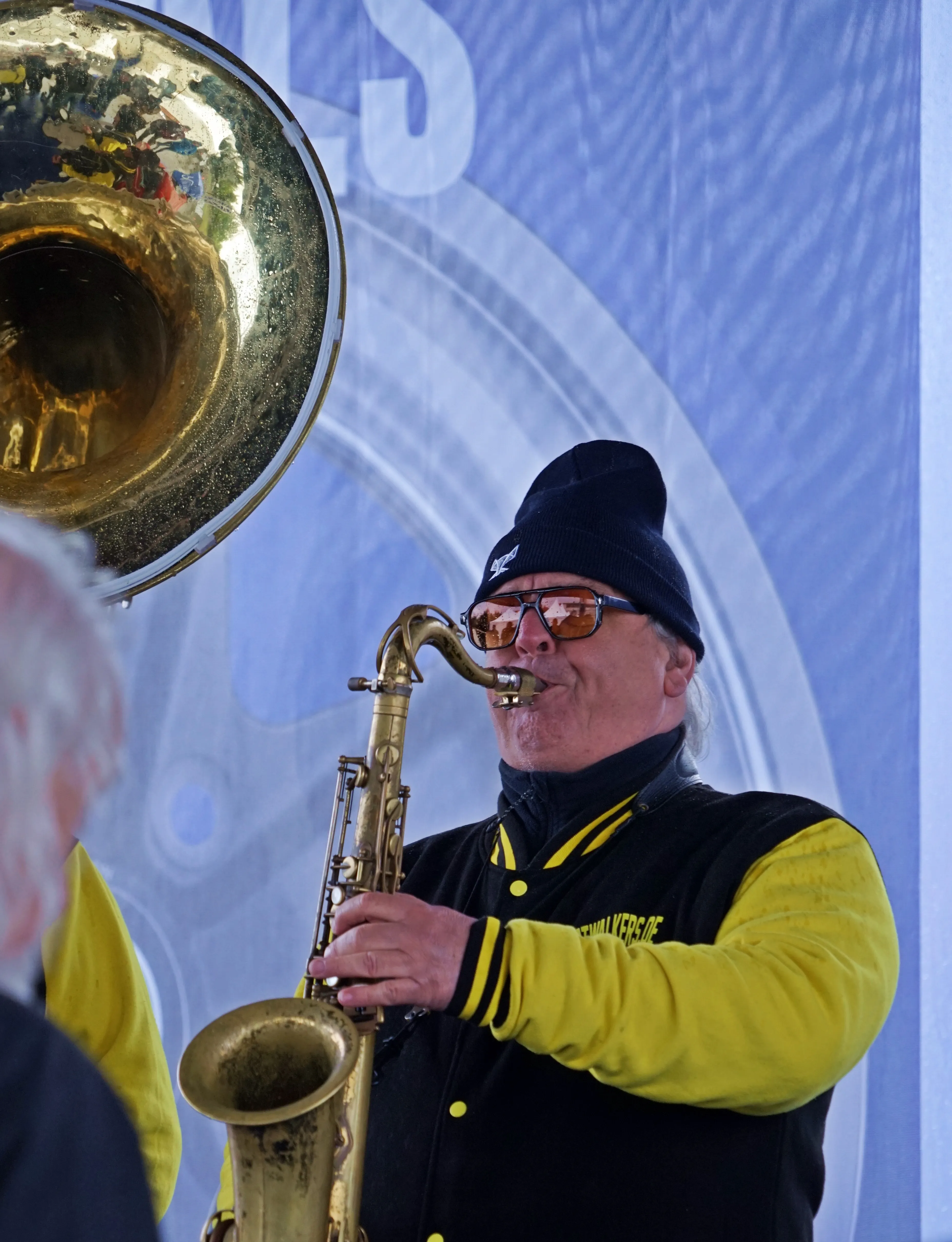 Ein Mann spielt Saxophon vor einer großen Posaune, während er eine Mütze und Sonnenbrille trägt. Im Hintergrund ist ein Banner mit einem abstrakten Muster zu sehen.
