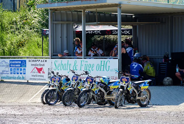 Mehrere Motoball-Fahrer stehen und sitzen vor ihren Motorrädern unter einem Pavillon mit Werbeaufdrucken. Im Hintergrund sind weitere Personen und ein Container zu sehen.