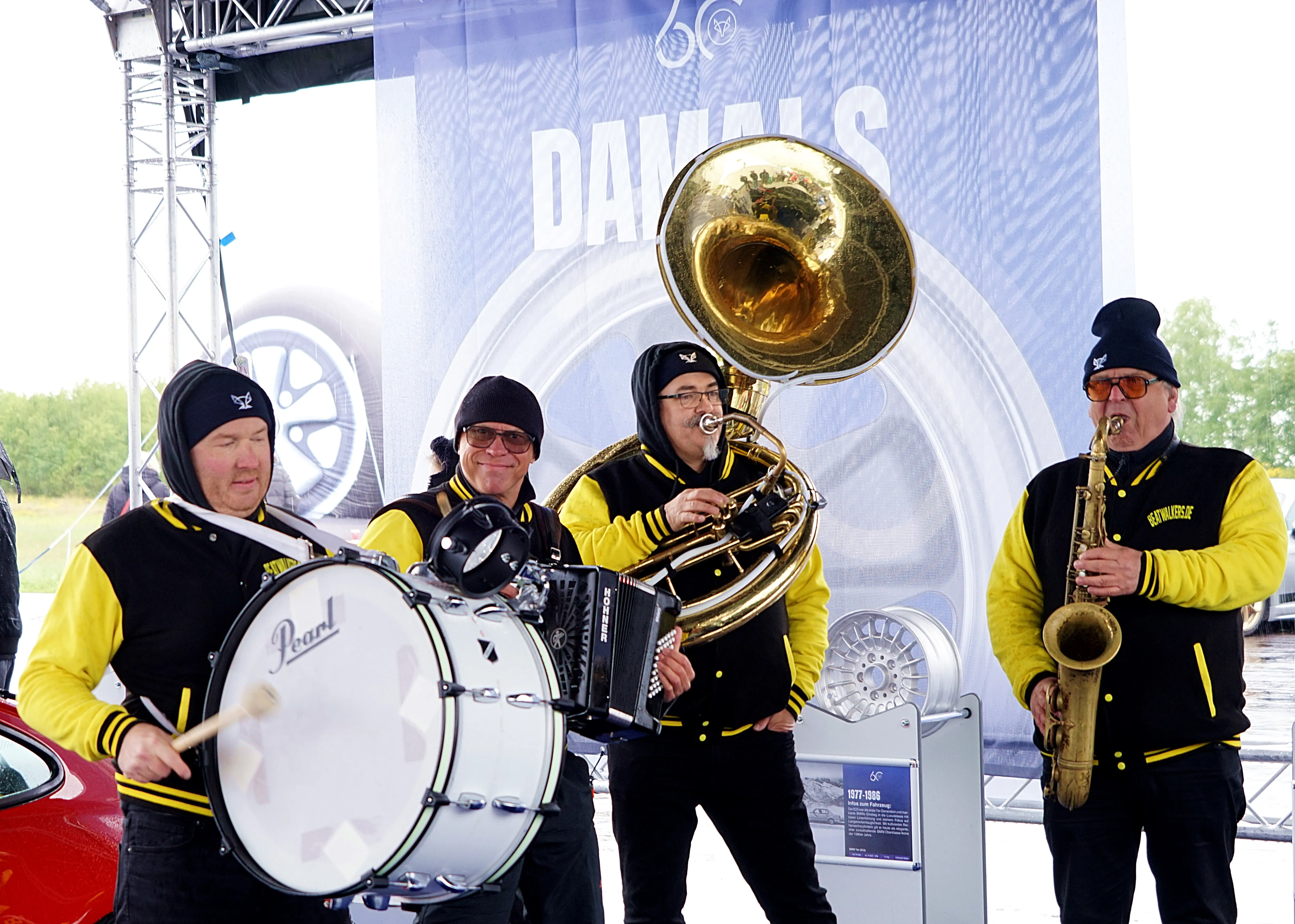 Vier Musiker spielen auf einem Freiluftgelände Blasinstrumente und Schlagzeug vor einer Bühne mit dem Schriftzug „DAWALS“.