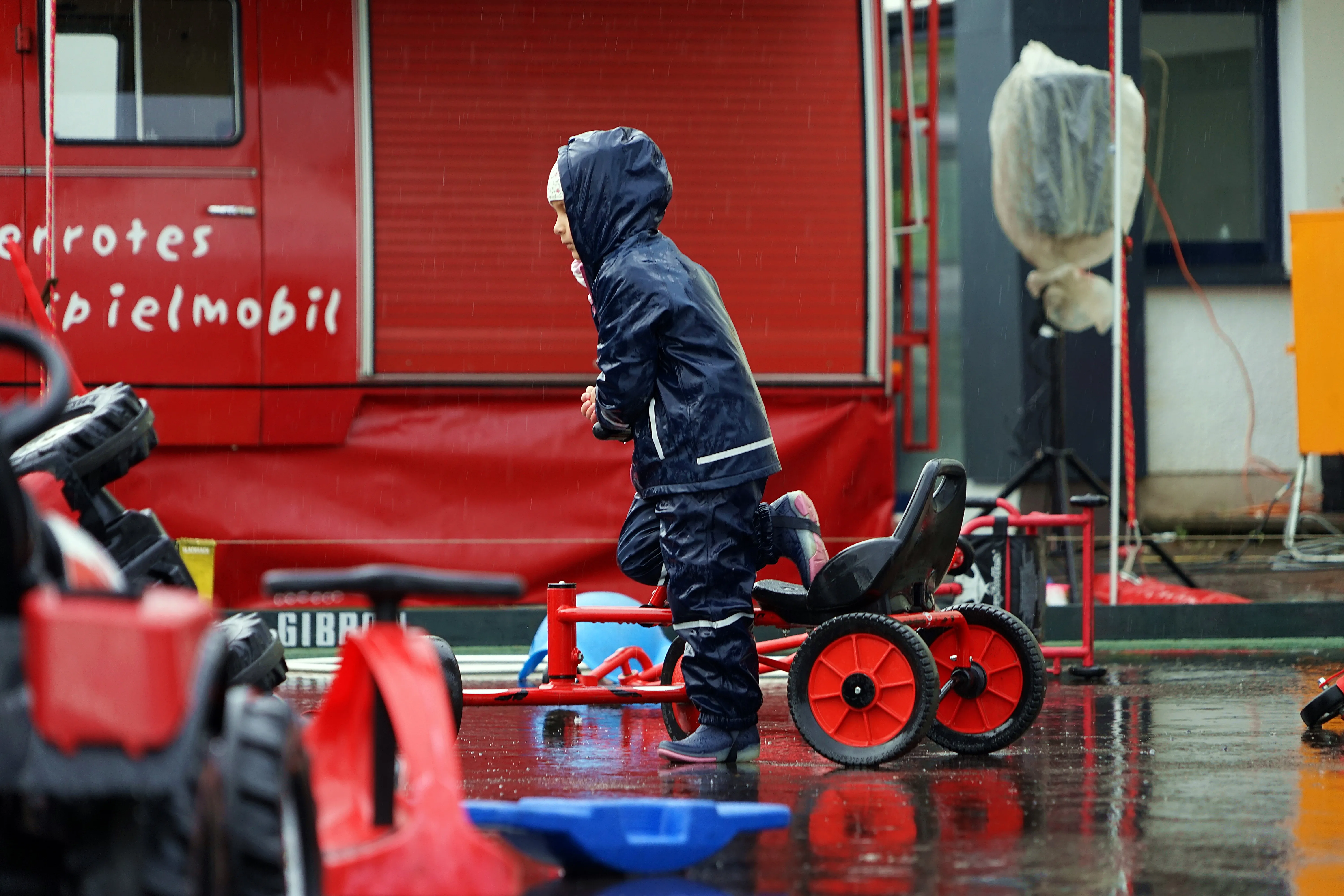 Ein Kind in Regenkleidung schiebt ein rotes Tretmobil auf einem nassen Platz. Im Hintergrund ein roter Transporter mit der Aufschrift „er rotes spielmobil“.