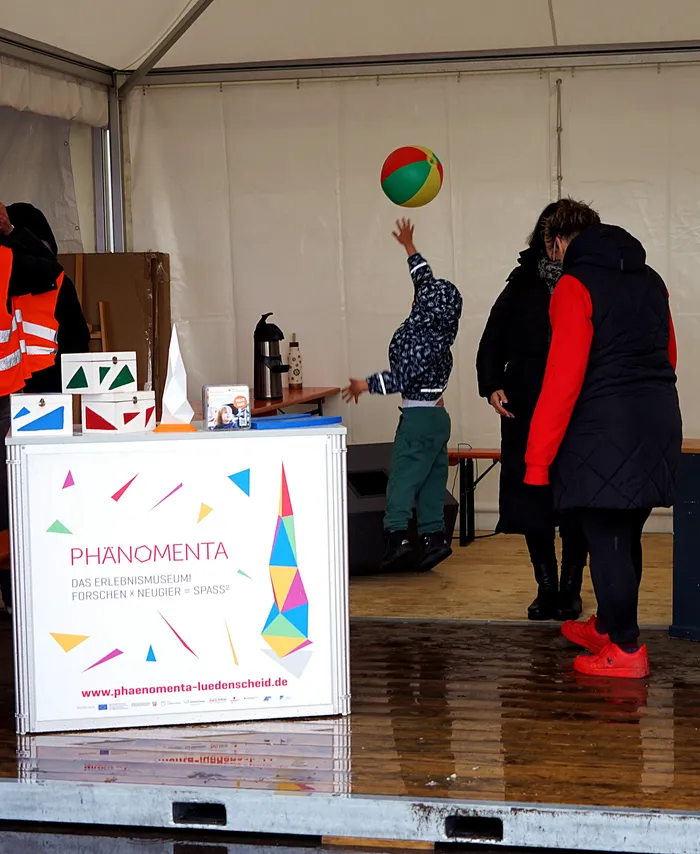 Ein Kind fängt einen bunten Ball vor einem Stand des Erlebnismuseums Phänomenta Luedenscheid. Im Hintergrund steht eine Person mit roter Jacke.