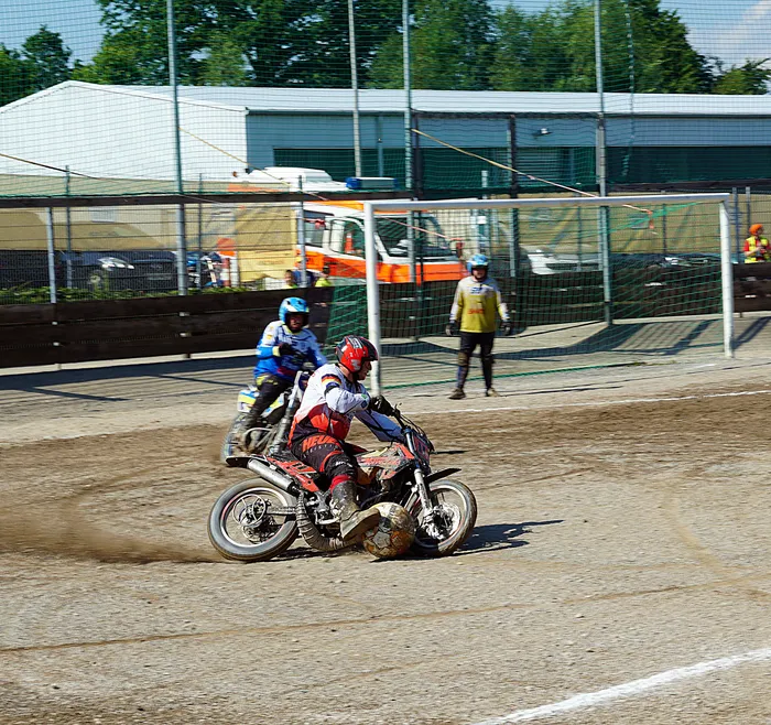 Zwei Motorradfahrer treten in einer sandigen Arena gegeneinander an, während im Hintergrund ein Fußballtor und Zuschauer zu sehen sind. Das Derby zwischen dem MSF Tornado Kierspe und dem MBC Kierspe ist in vollem Gange.