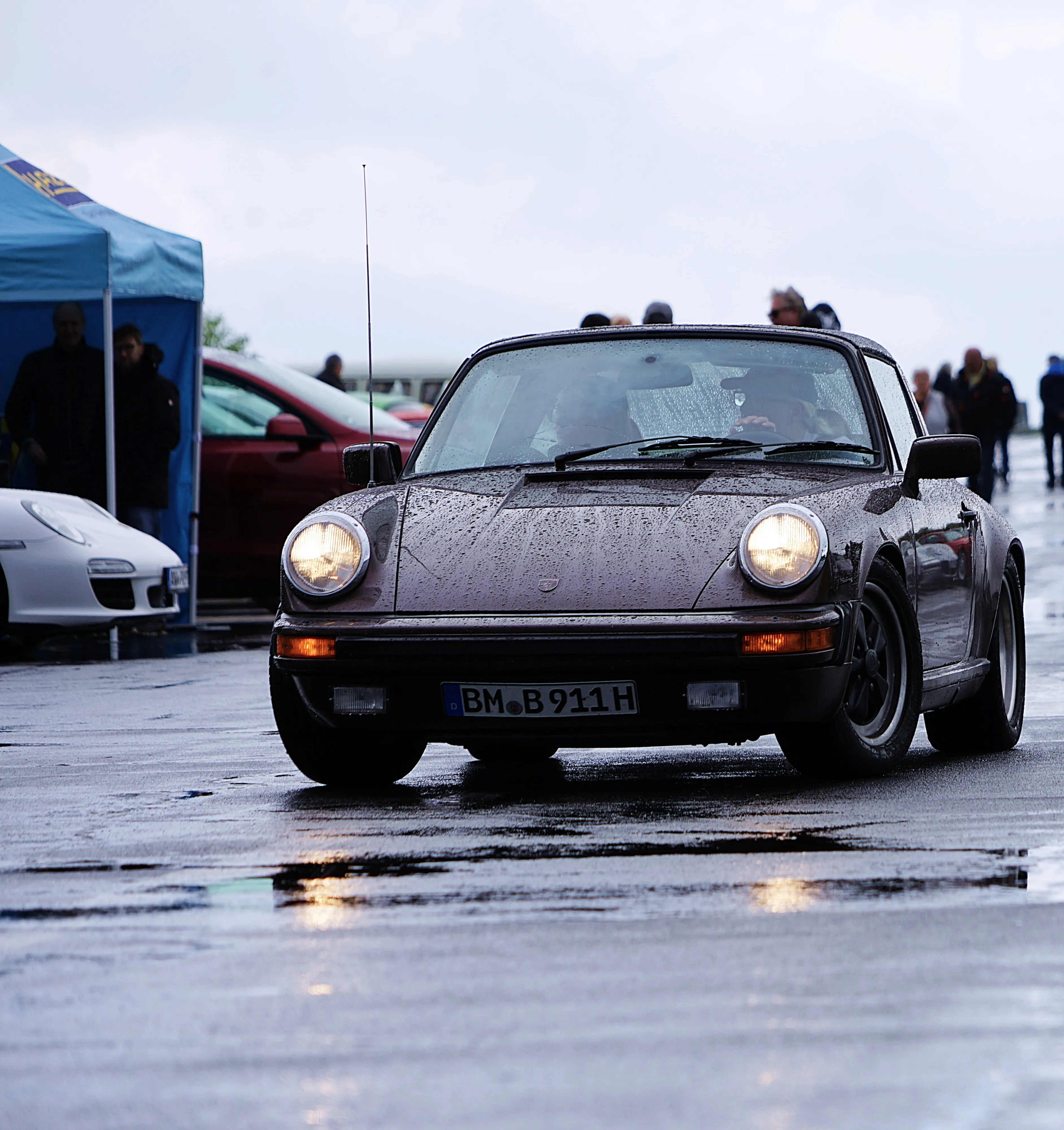 Dunkler Porsche 911 mit Fuchsfelgen steht auf nasser Fahrbahn, Kennzeichen BM-B 911H ist erkennbar. Im Hintergrund weitere Fahrzeuge und ein Zelt.