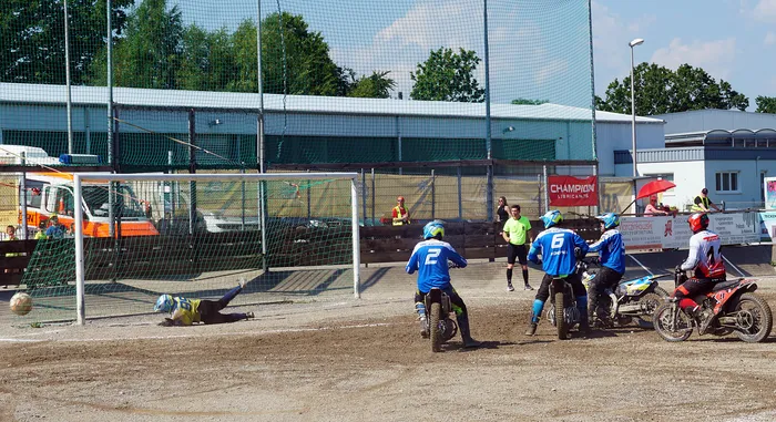 Motoballspieler auf Motorrädern kämpfen um den Ball vor dem Tor, während ein Torwart versucht, den Angriff zu vereiteln. Im Hintergrund eine Sportanlage mit Zuschauern und Werbebanden.