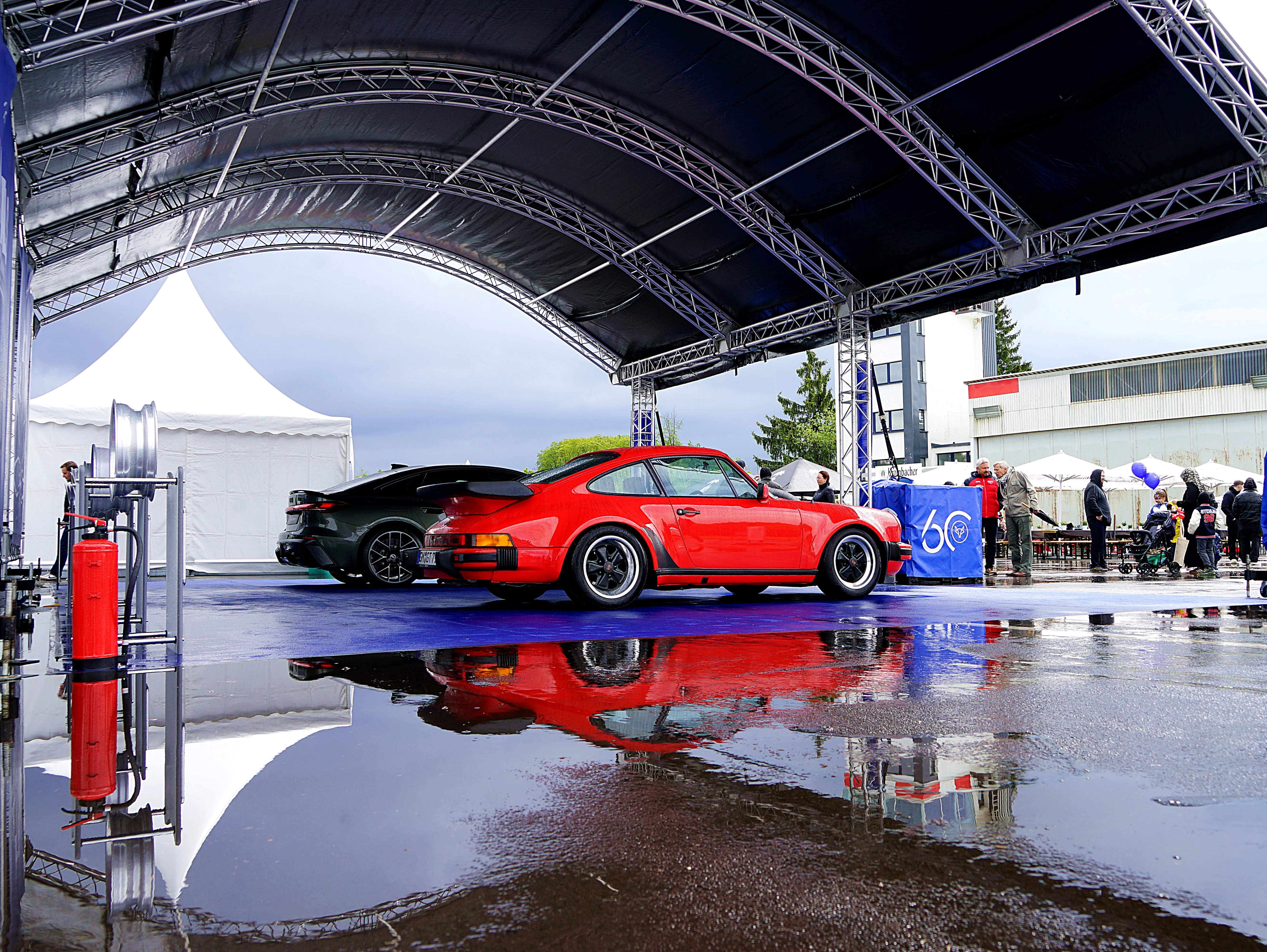 Roter Porsche 911 steht auf einer nassen Fläche unter einem großen Zeltdach, im Hintergrund weitere Fahrzeuge und Besucher.