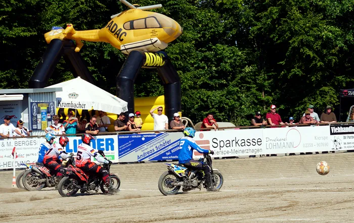 Motorradfahrer treten bei einem Motoball-Spiel gegeneinander an, während Zuschauer im Hintergrund zusehen. Im Hintergrund befindet sich ein großes, aufblasbares Luftschiff mit dem ADAC-Logo.