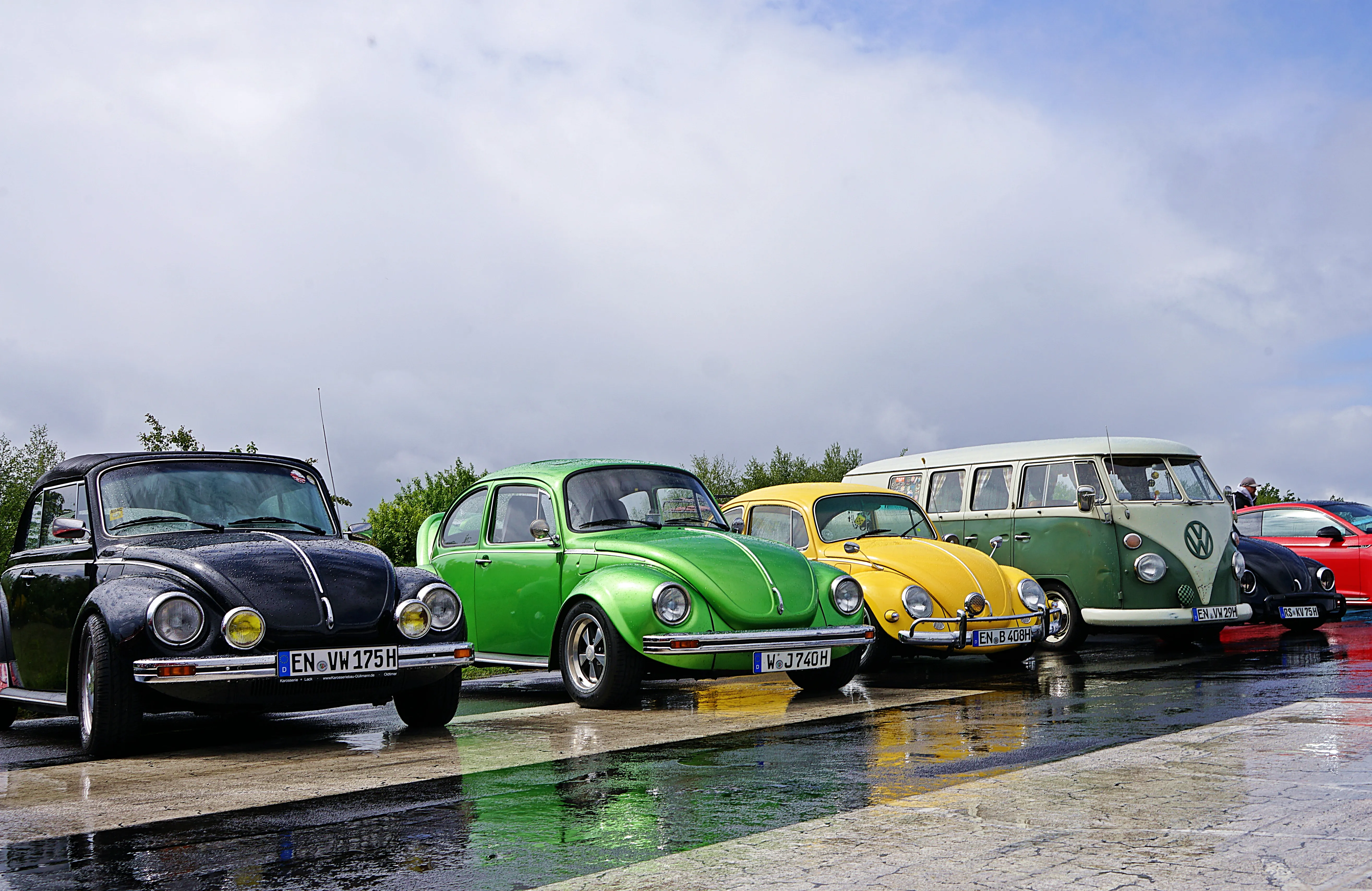 Reihe klassischer Volkswagen Käfer und ein Bulli stehen auf einem nassen Pflaster. Die Fahrzeuge sind in verschiedenen Farben lackiert und tragen Fuchsfelgen.