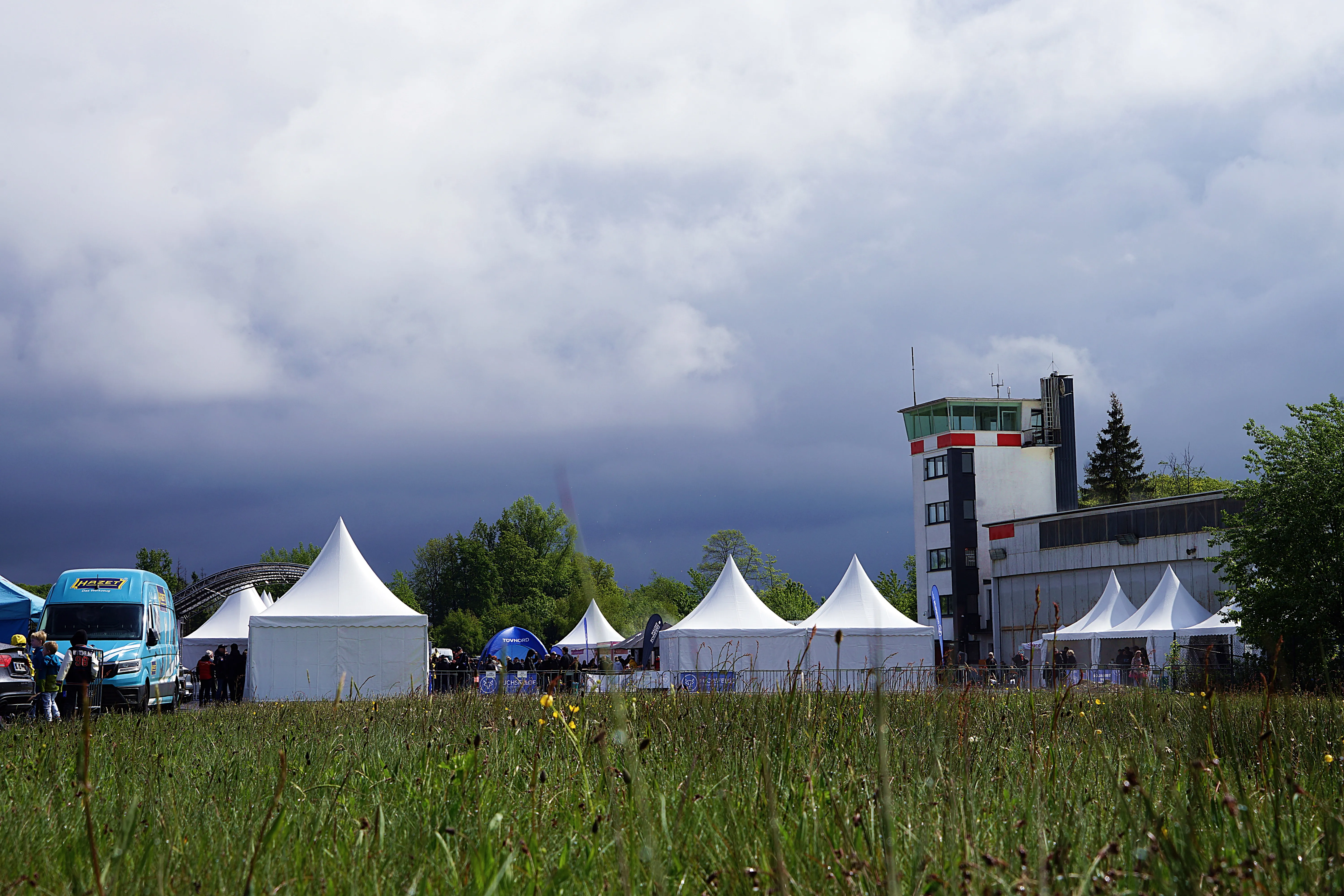 Ein Feld mit Wildblumen im Vordergrund, dahinter mehrere weiße Zelte und ein Kontrollturm unter einem bewölkten Himmel beim Fuchsfelgen-Treffen.