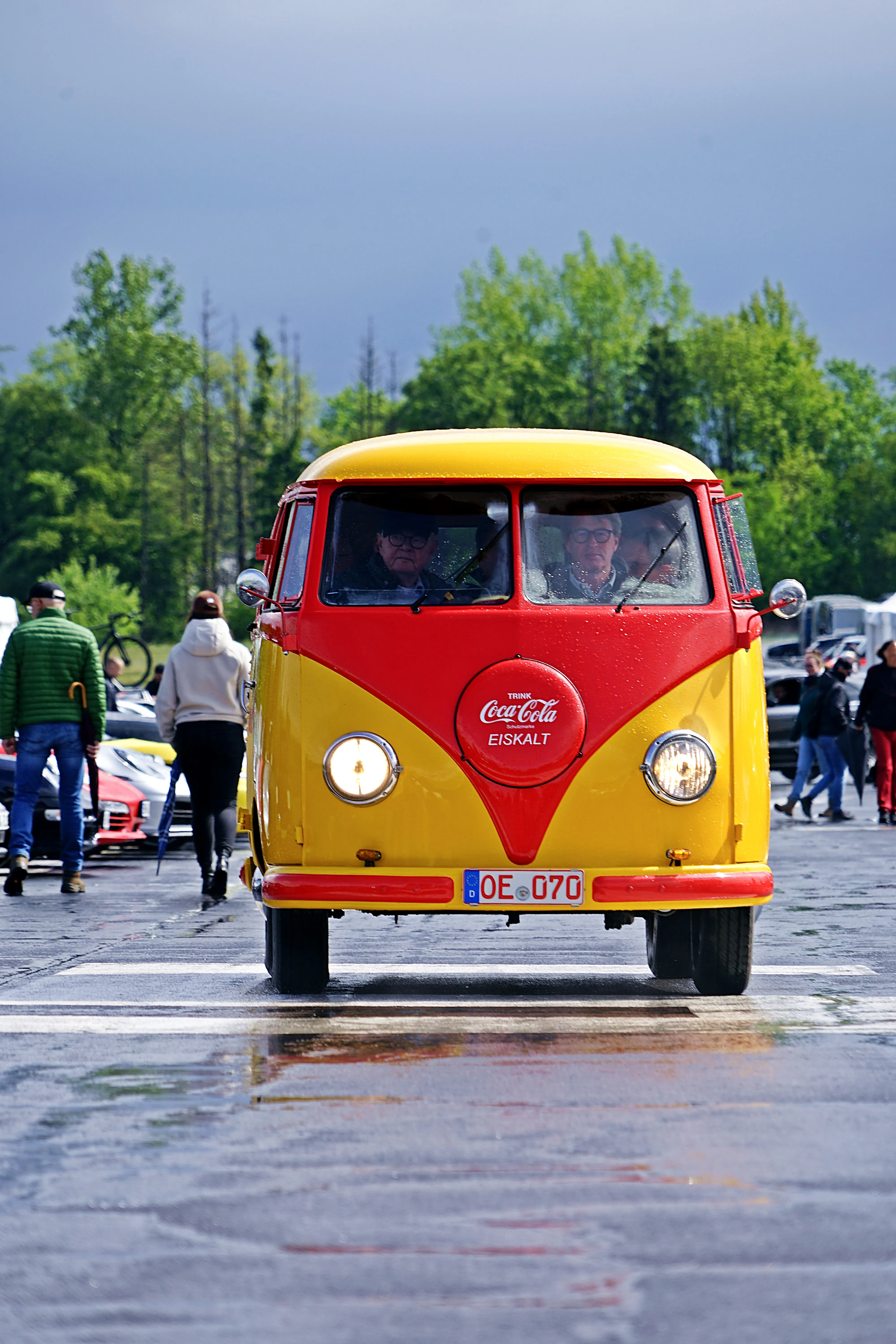 Gelb-roter VW-Bus mit Coca-Cola-Emblem fährt auf nasser Straße vor Zuschauern, Fuchsfelgen-Treffen 2026.