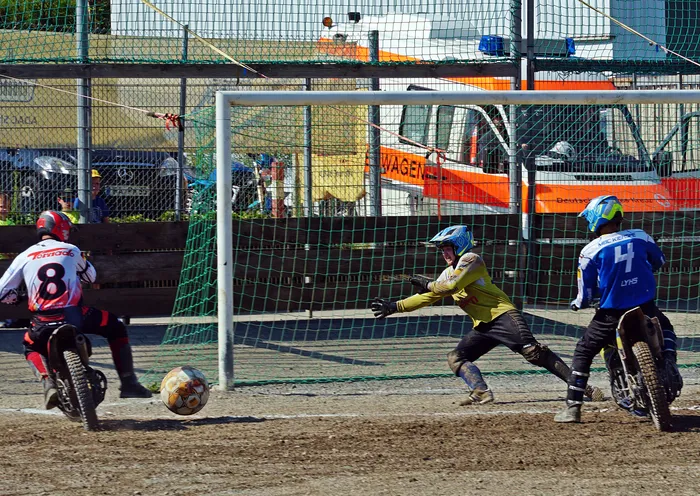 Ein Motoball-Spieler schießt auf das Tor, während der Torwart versucht, den Ball abzuwehren. Im Hintergrund sind weitere Spieler und eine Absperrung zu sehen.