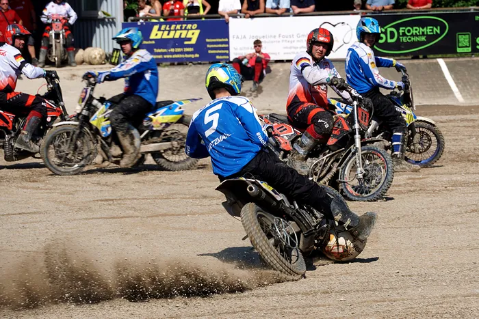 Motoballspieler auf Motorrädern während eines Spiels, wobei ein Spieler in blauem Trikot mit der Nummer 5 in den Vordergrund fährt und versucht, den Ball zu erobern. Im Hintergrund weitere Spieler und Zuschauer.