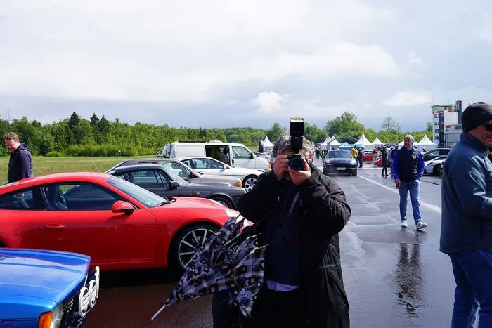 Ein Fotograf hält eine Kamera mit Blitz vor einer Reihe geparkter Sportwagen, während im Hintergrund Besucher auf einem Gelände unterwegs sind.