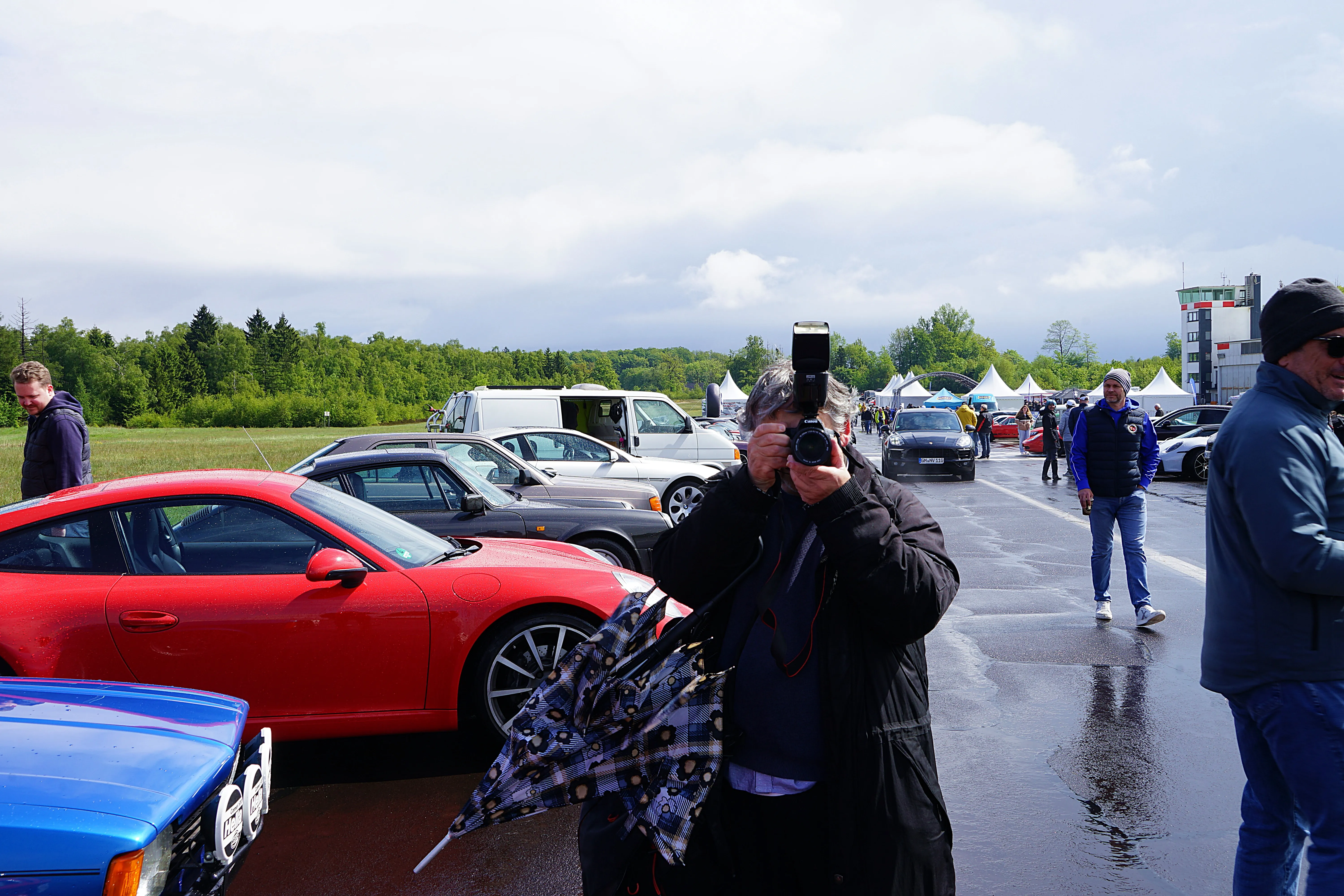Ein Fotograf hält eine Kamera mit Blitz vor einer Reihe geparkter Sportwagen, während im Hintergrund Besucher auf einem Gelände unterwegs sind.
