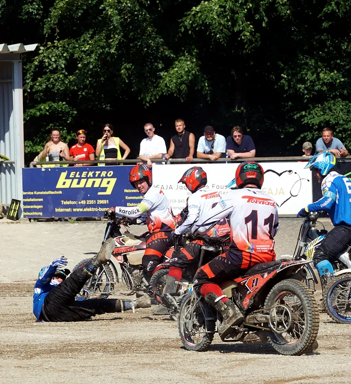 Mehrere Motoballspieler auf Motorrädern starten ein Rennen auf einer Sandbahn, umgeben von Zuschauern und Werbebannern.