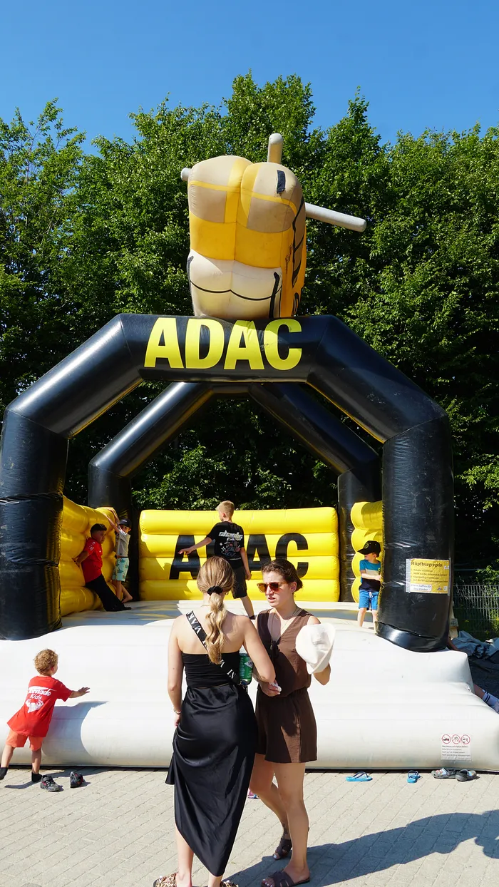 Aufblasbare ADAC-Burg in Form eines Motorradhelms steht im Vordergrund, vor der zwei Frauen stehen und sich unterhalten. Im Hintergrund sind weitere Personen und ein Kind zu sehen, die sich in der Nähe der Burg aufhalten.