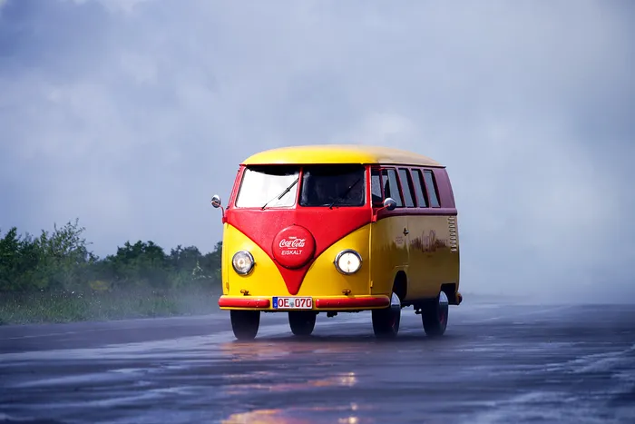 Gelb-roter VW-Bus mit Coca-Cola-Werbung fährt auf nasser Straße vor bewölktem Himmel.