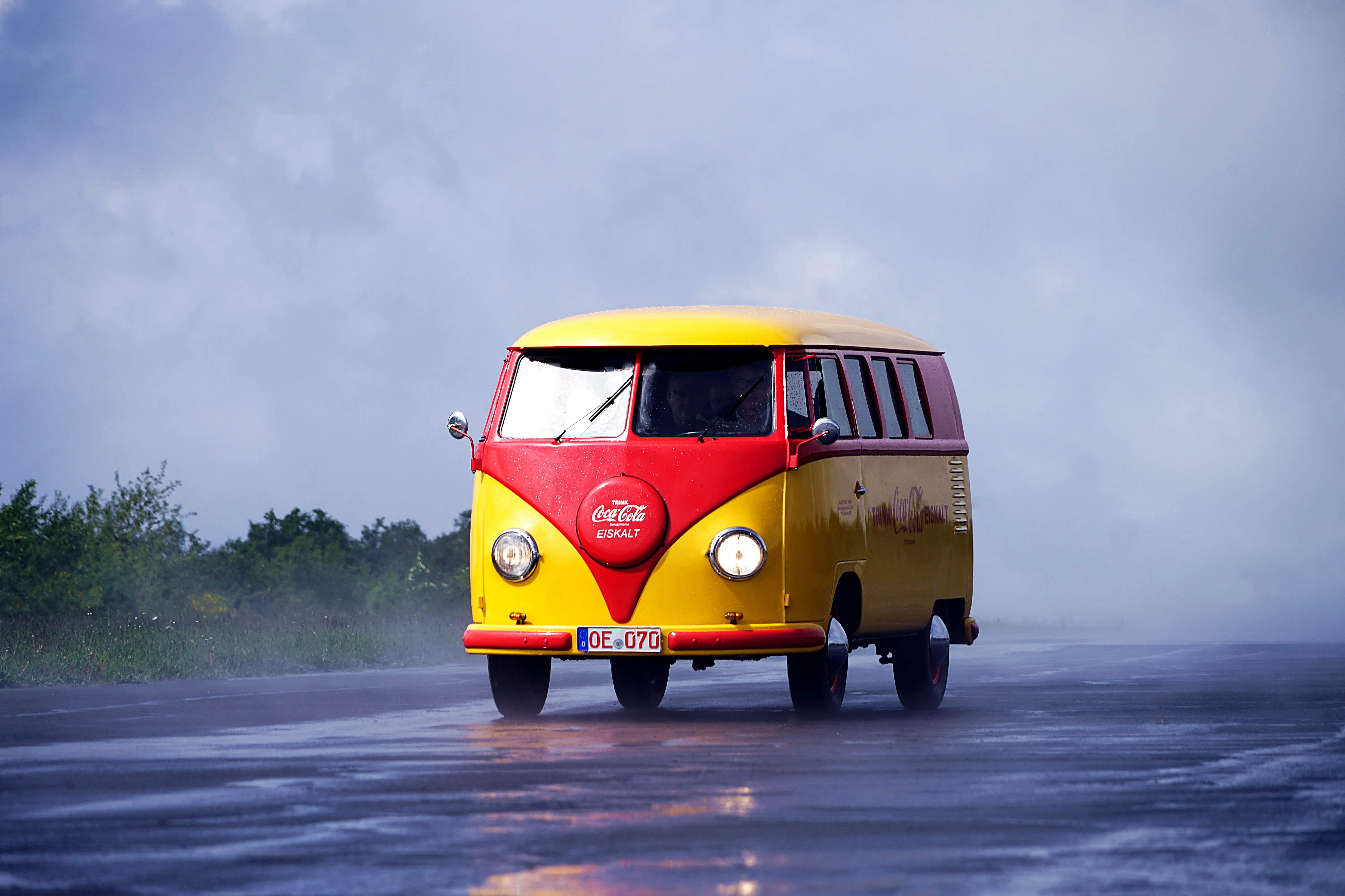 Gelb-roter VW-Bus mit Coca-Cola-Werbung fährt auf nasser Straße vor bewölktem Himmel.