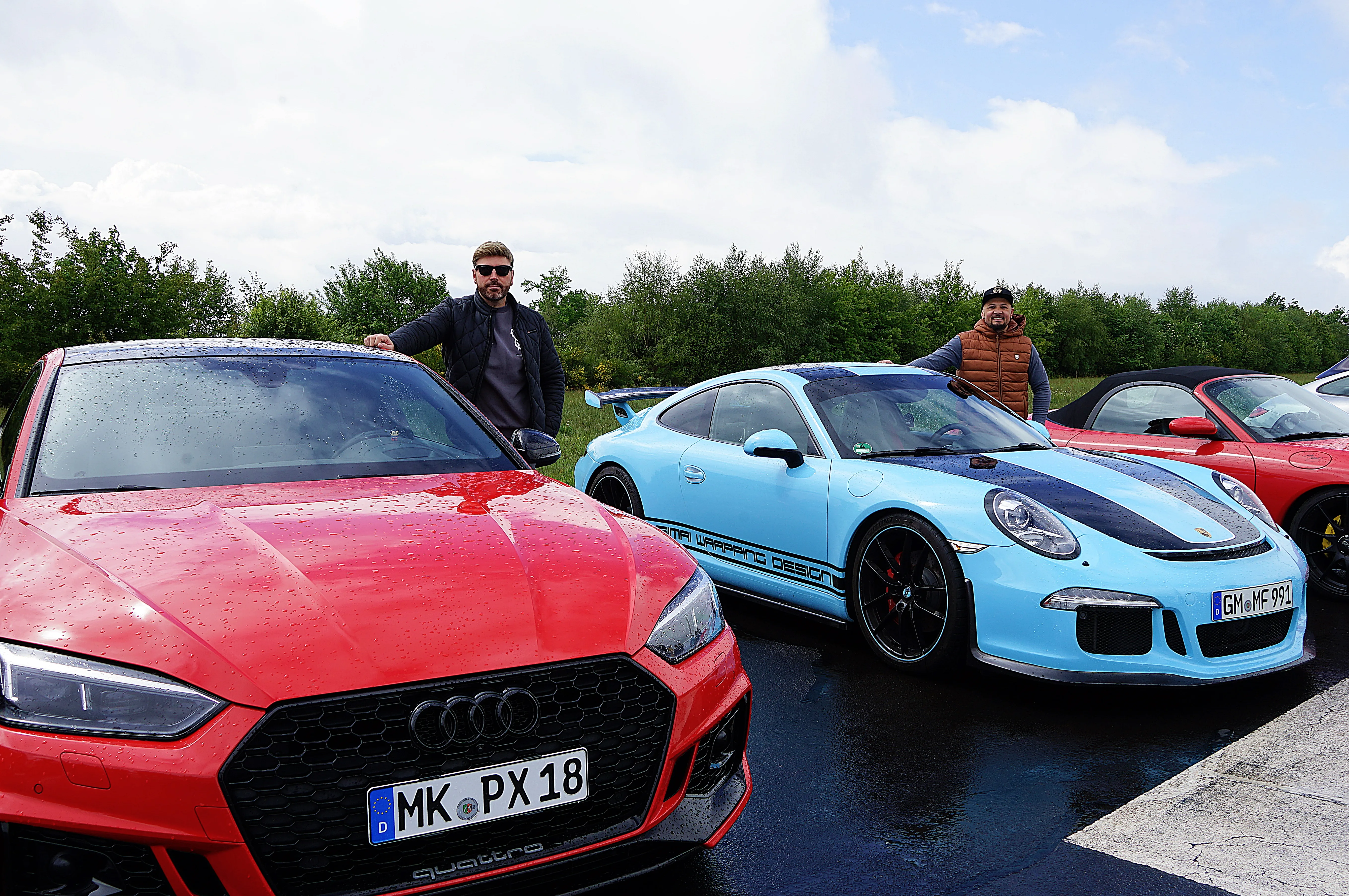 Zwei Männer stehen vor ihren Sportwagen auf einem Parkplatz. Links ein roter Audi RS3, rechts ein hellblauer Porsche 911 mit schwarzem Dach.