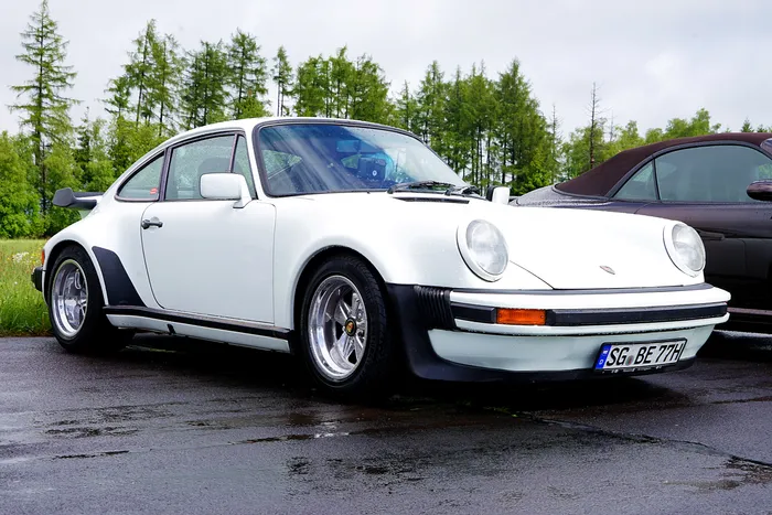 Weißer Porsche 911 mit Fuchsfelgen steht auf einem nassen Parkplatz, Bäume im Hintergrund.