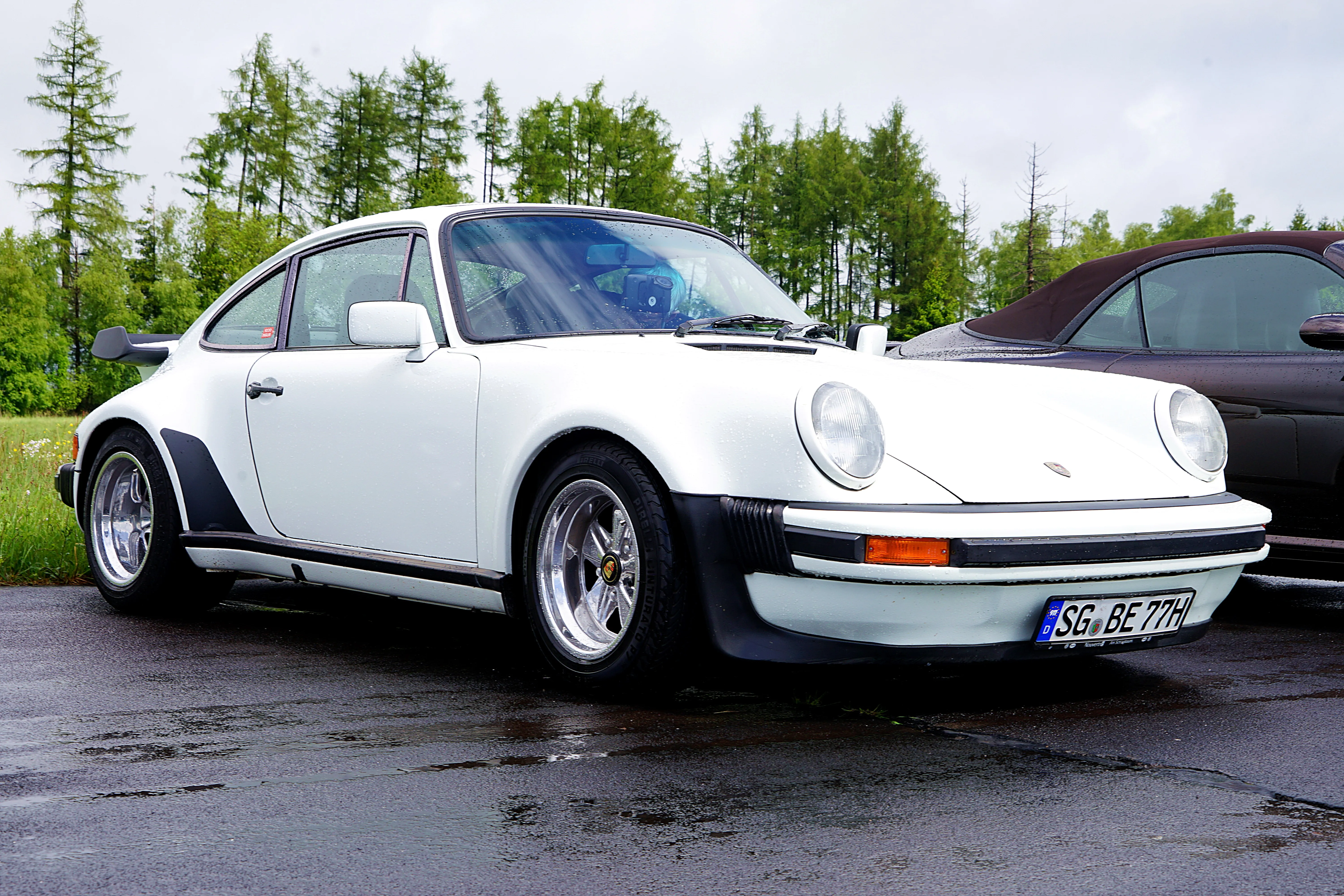 Weißer Porsche 911 mit Fuchsfelgen steht auf einem nassen Parkplatz, Bäume im Hintergrund.