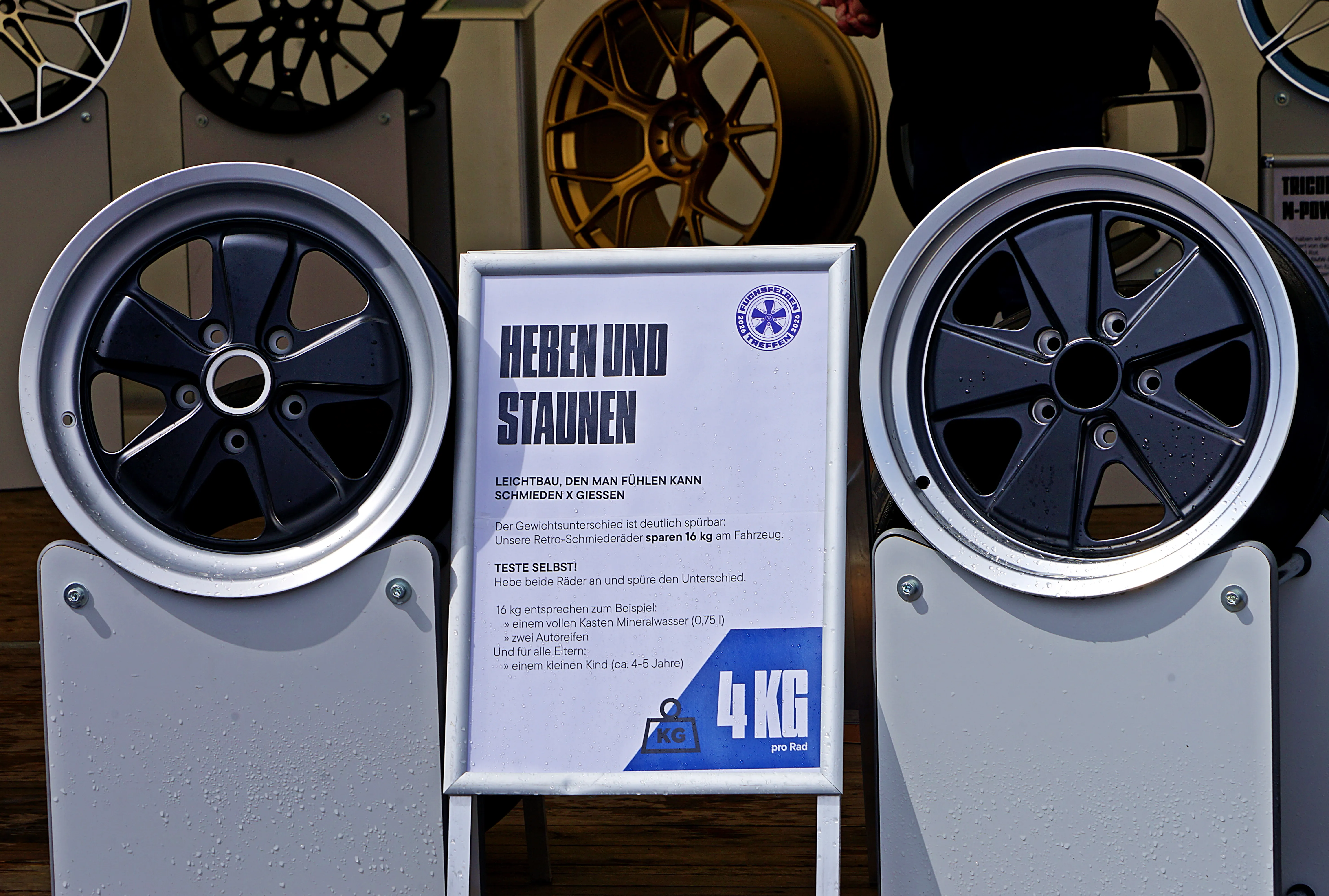 Zwei schwarze Fuchsfelgen stehen vor einem Werbeschild mit dem Aufdruck „Heben und Staunen“ und dem Hinweis auf ein Gewicht von 4 kg pro Rad.