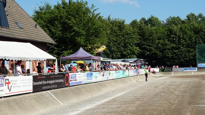 Zuschauer beobachten ein Motoball-Spiel auf einer Betonbahn, auf der zwei Spieler mit Motorrädern gegeneinander antreten. Im Hintergrund sind Gebäude und Bäume zu sehen.